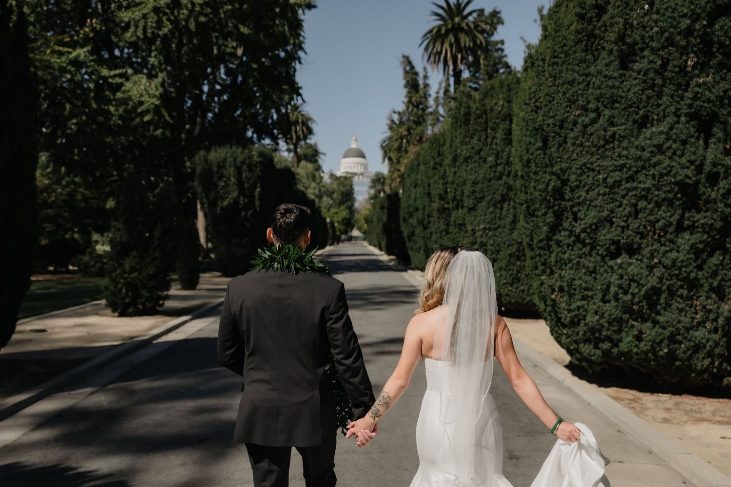 Bride in a white gown and veil holds hands with a groom in a dark suit while walking down a tree-lined path toward a distant capitol dome.