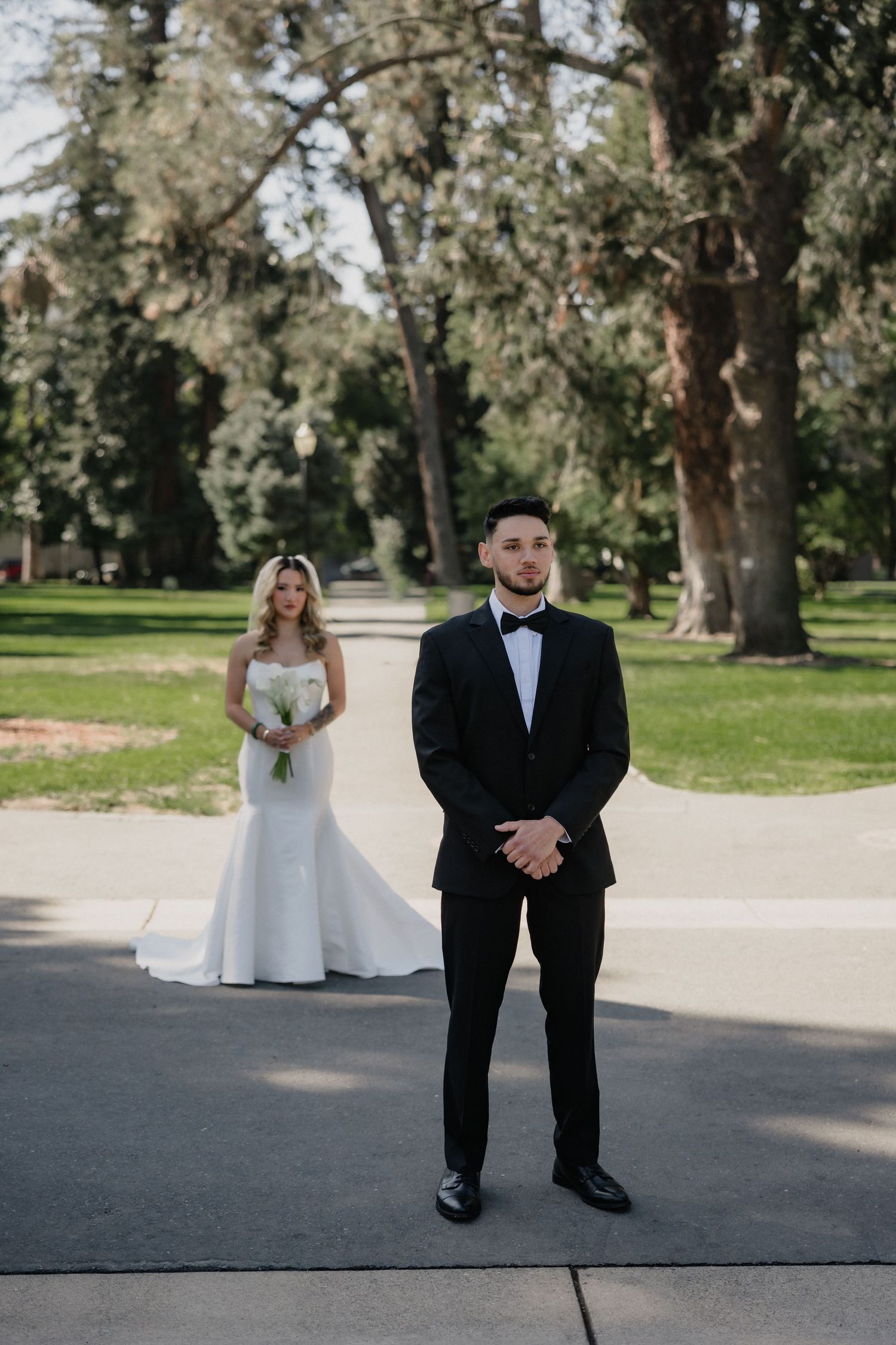 Groom in a black tuxedo and bow tie stands on a park path while the bride in a white gown waits behind him for a first look.
