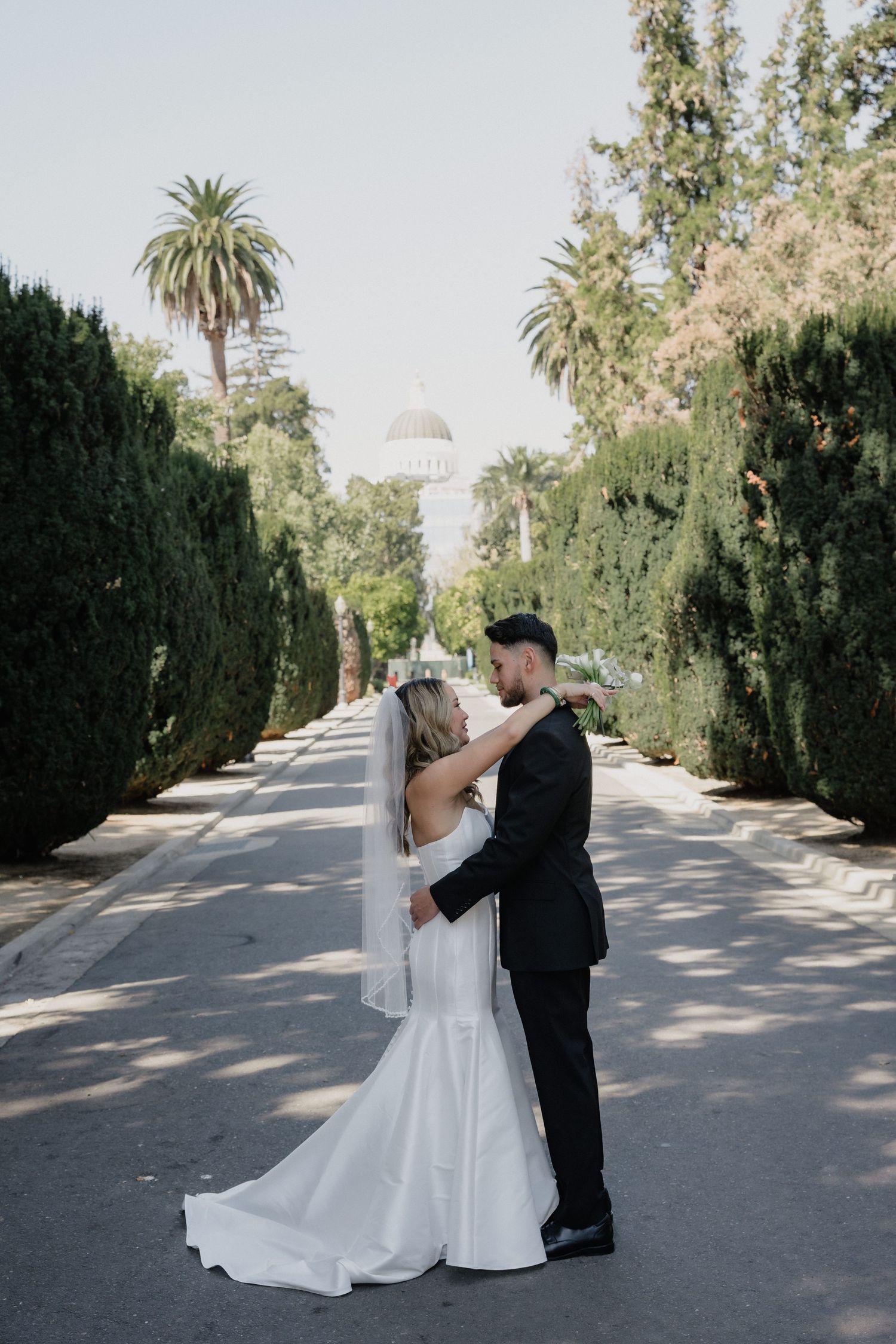 Bride in a white mermaid gown and veil embraces her groom in a black suit on a tree-lined path with a domed building behind.