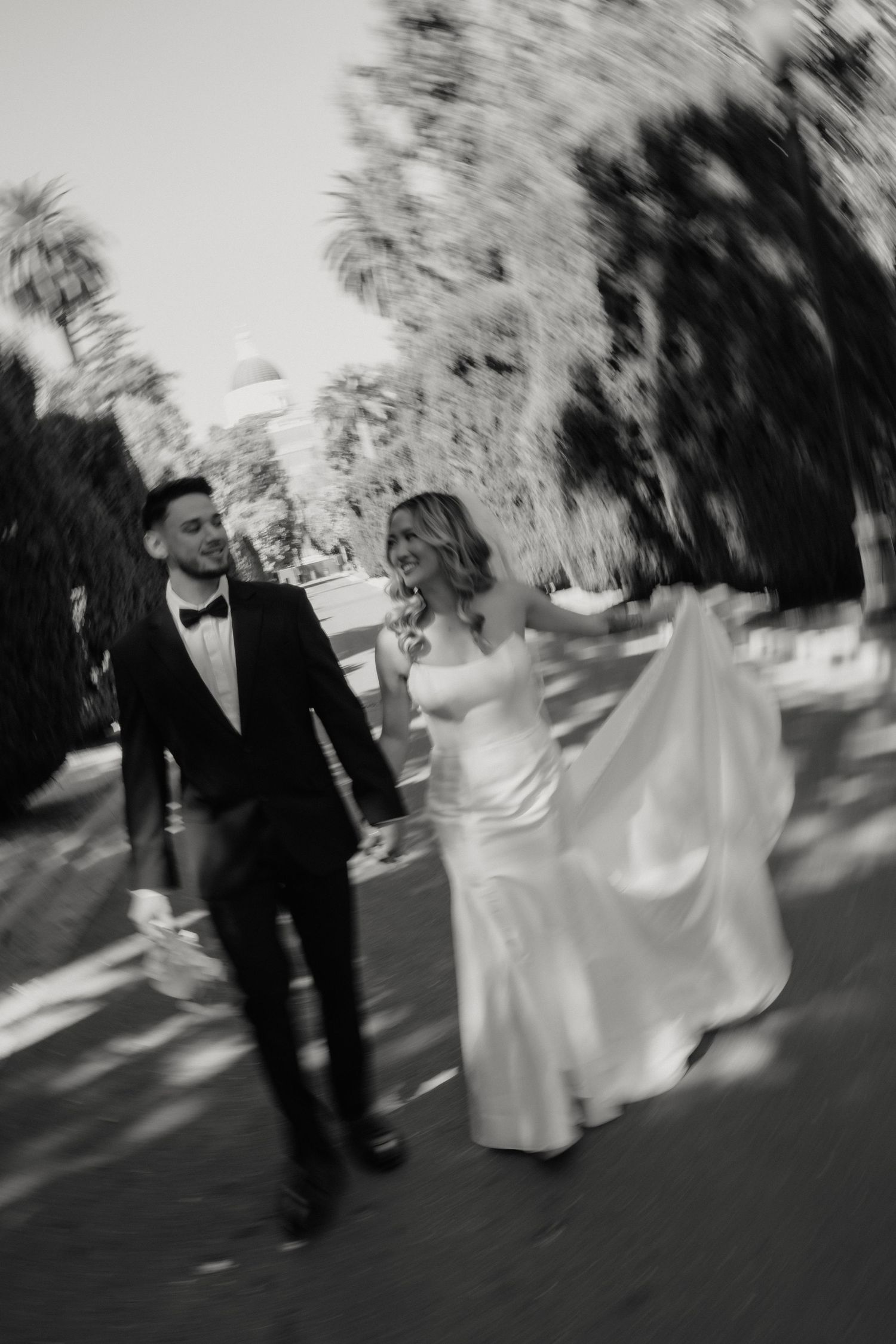 Black and white photo of a bride in a satin gown and groom in a tuxedo walking hand in hand along a tree-lined path.