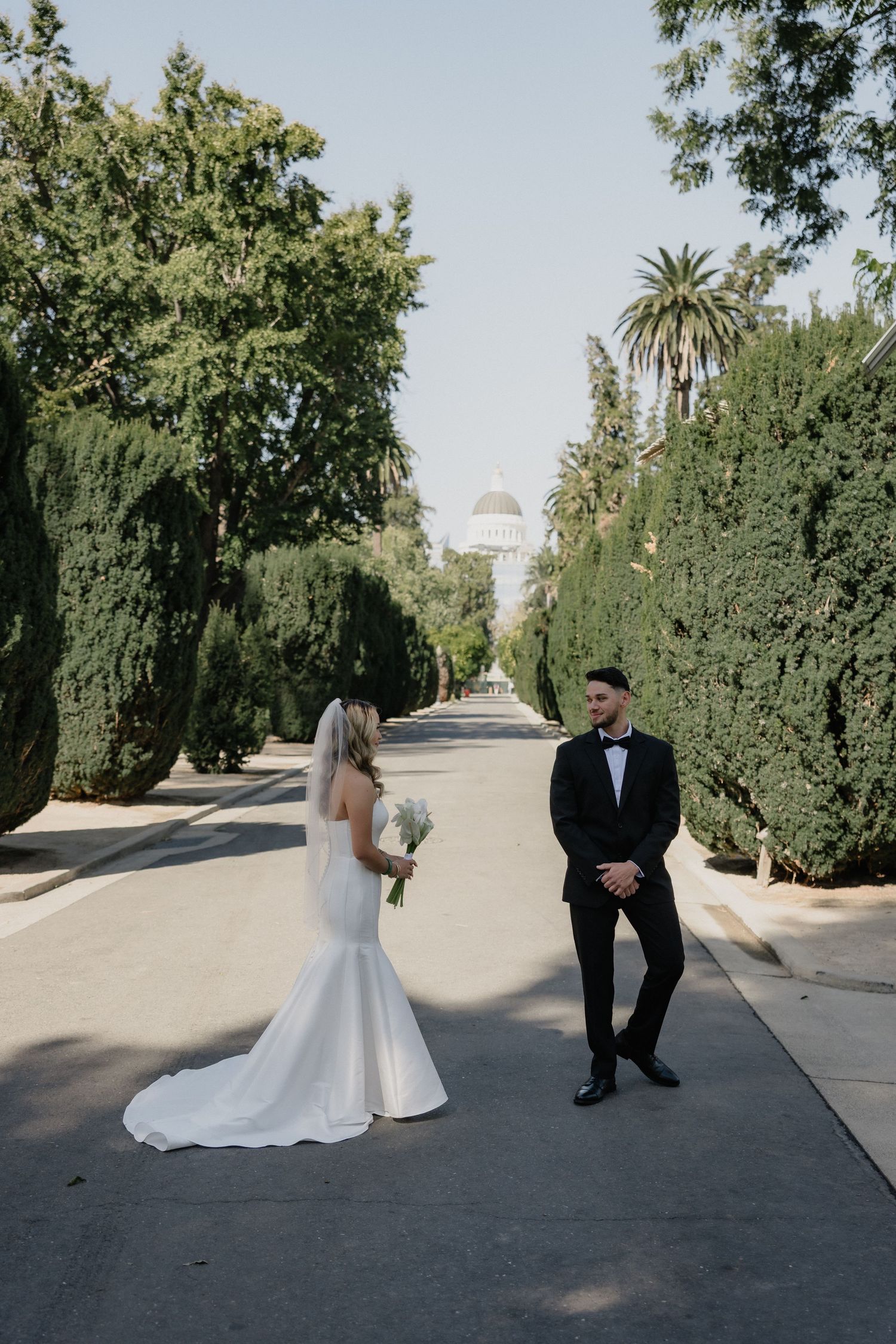 Bride in a white gown holding a bouquet faces a groom in a black tuxedo on a tree-lined road leading to a domed capitol building.