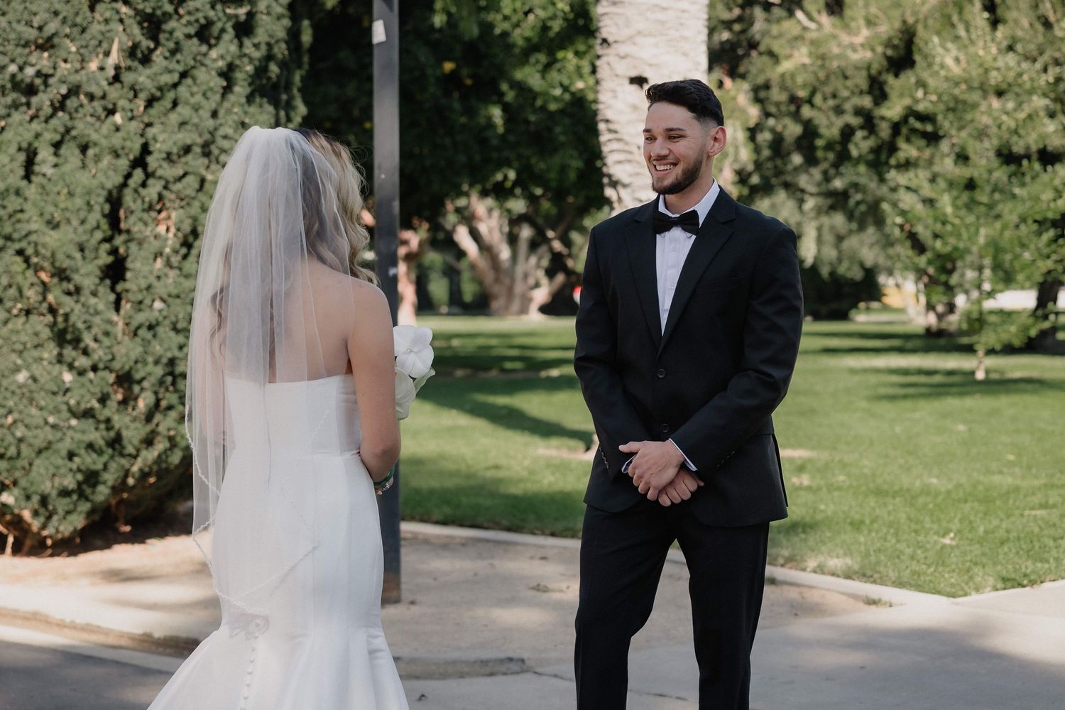 Groom in a black tuxedo and bow tie smiles at a bride in a white gown and veil during a first look in a sunny, tree-filled park.