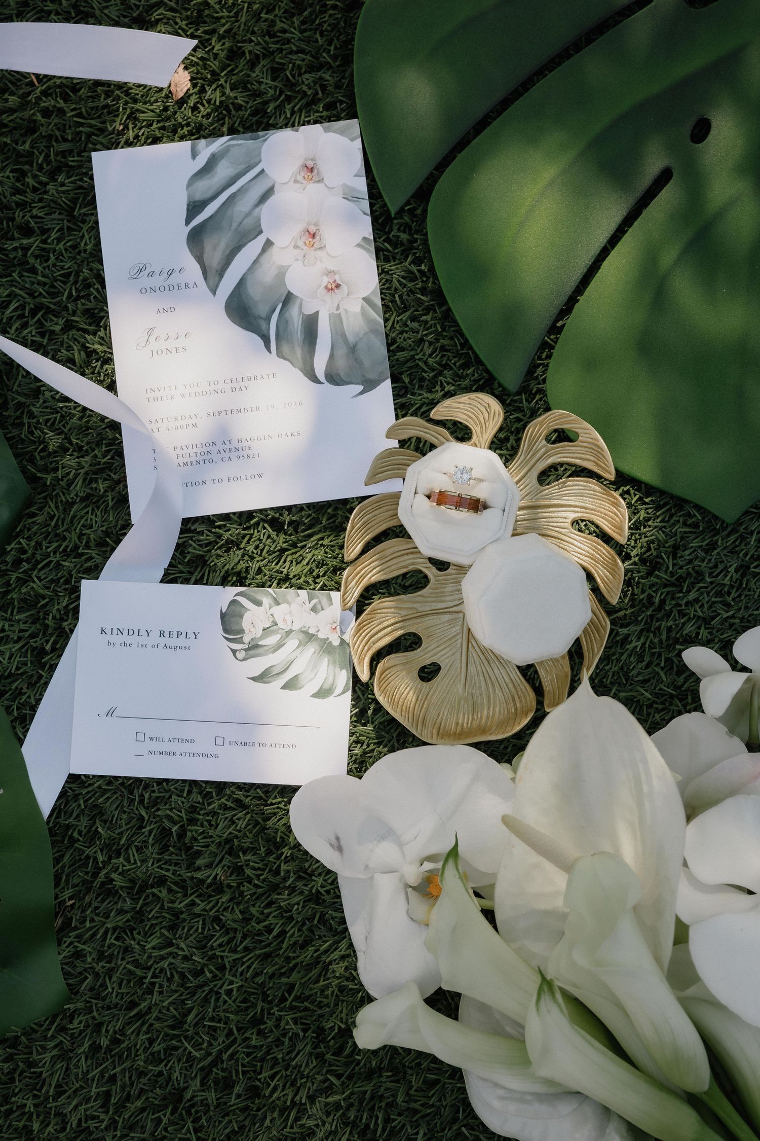 Tropical wedding invitation and RSVP card with a gold monstera leaf ring dish, white calla lilies, and green palm leaves on grass.