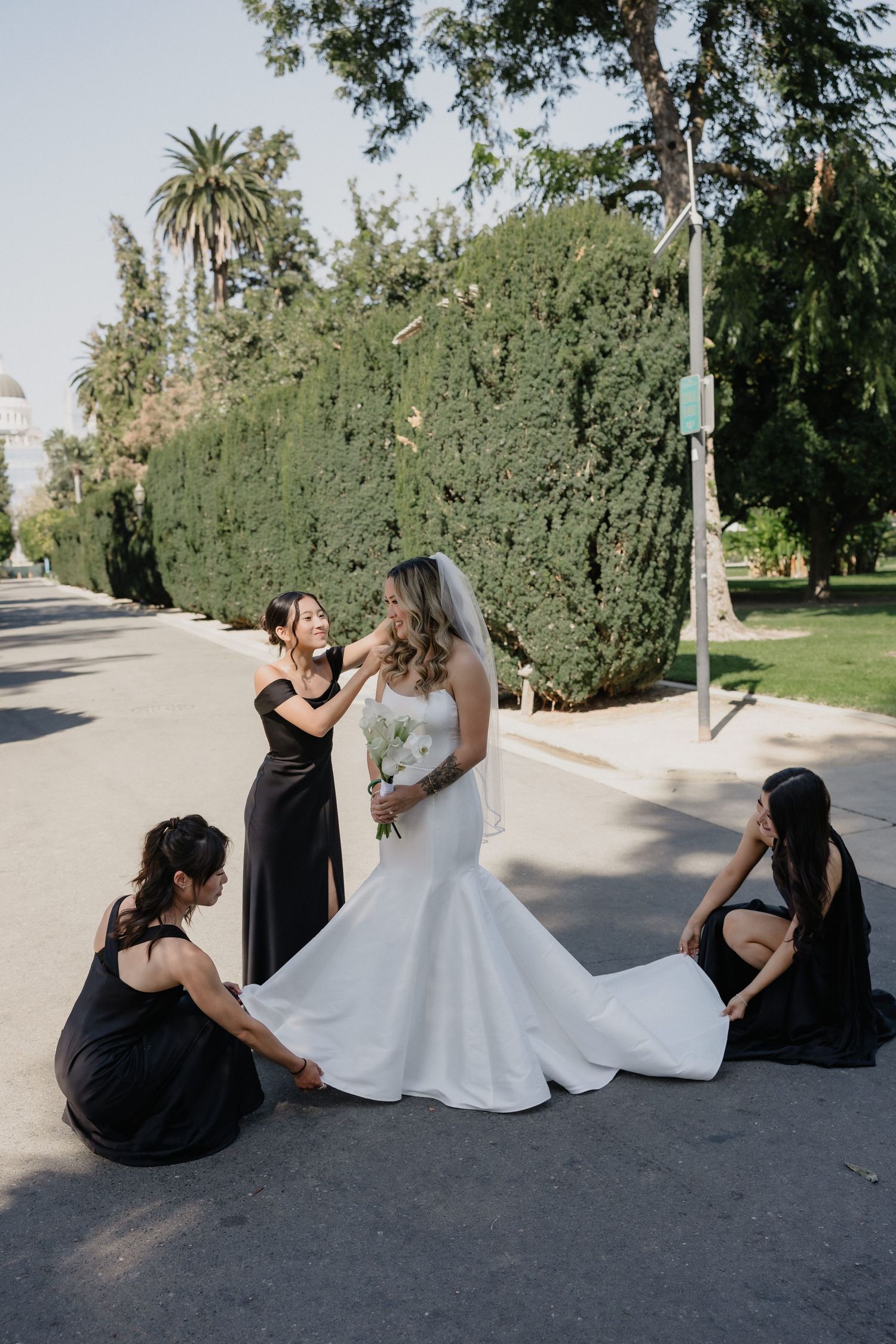 Three bridesmaids in black dresses adjust the white mermaid-style wedding gown and veil of a bride holding a bouquet on a tree-lined street.