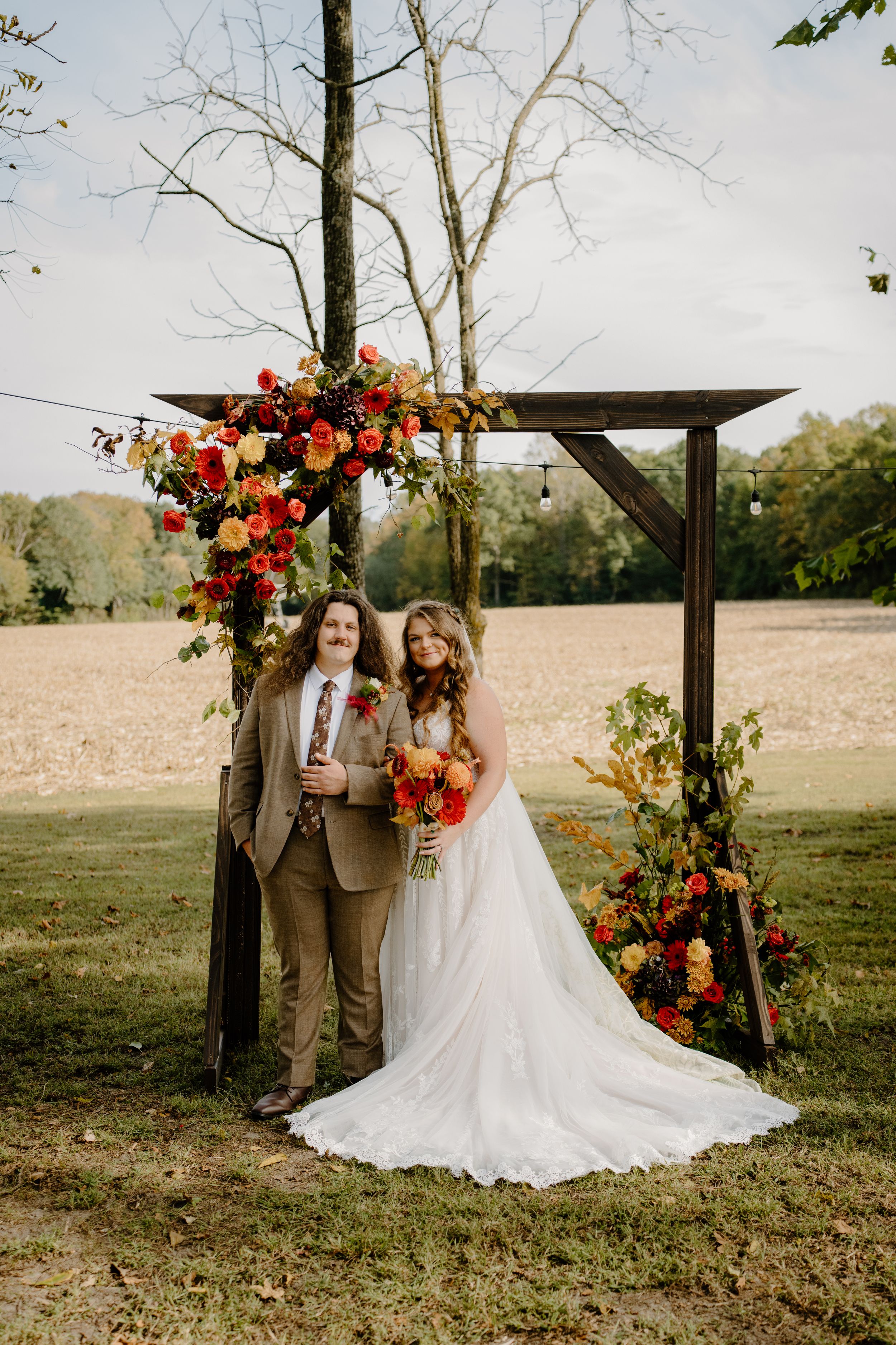 Rustic outdoor wedding ceremony setup with wooden arch decorated in vibrant fall flowers and autumn foliage in a pastoral field.