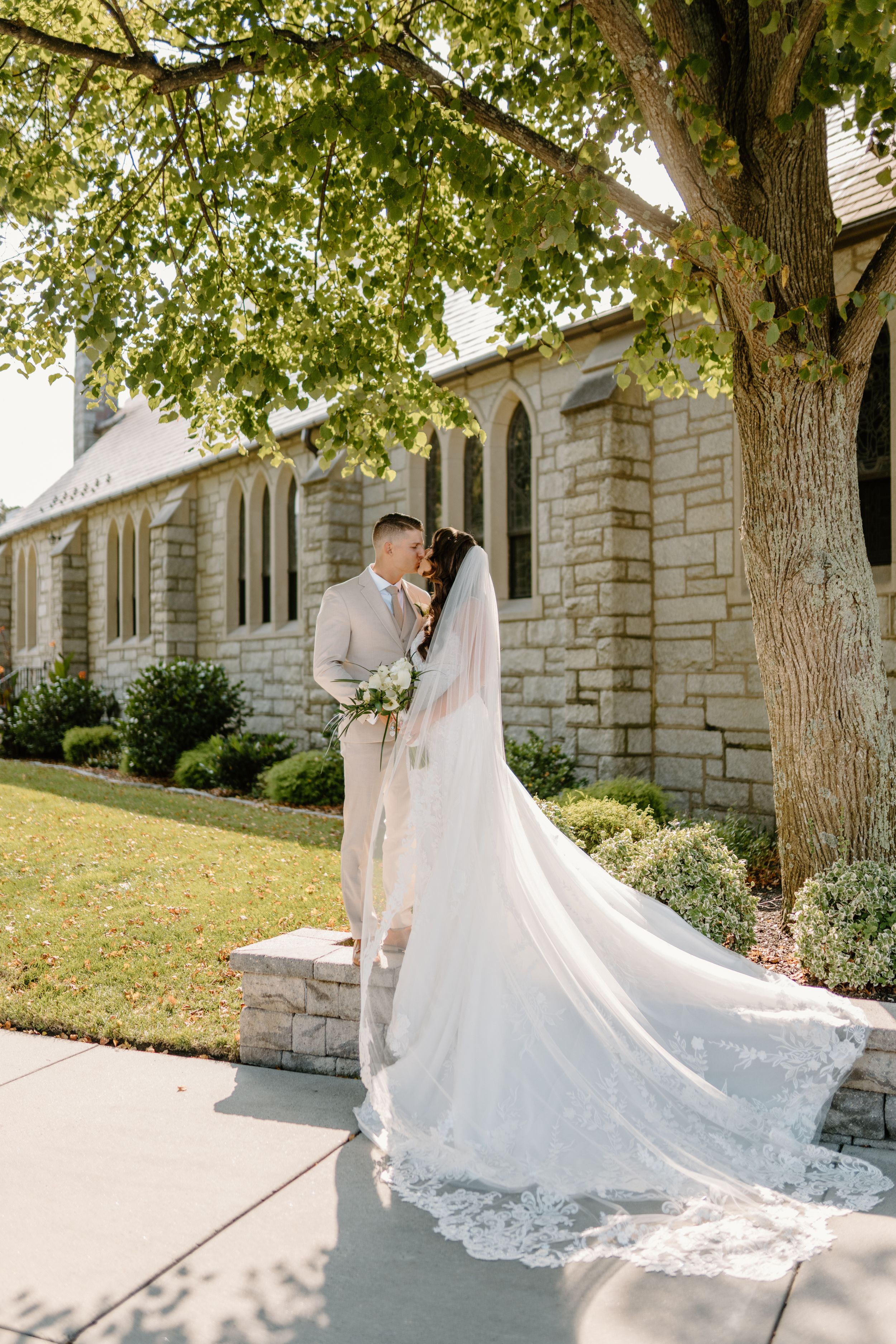 Wedding couple embracing outside a stone church building under a large tree on a sunny day.