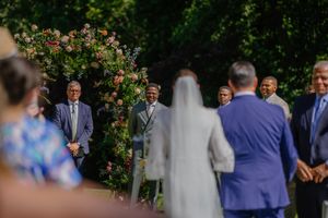 Outdoor wedding ceremony with bride and groom at floral arch surrounded by guests in a lush garden setting.