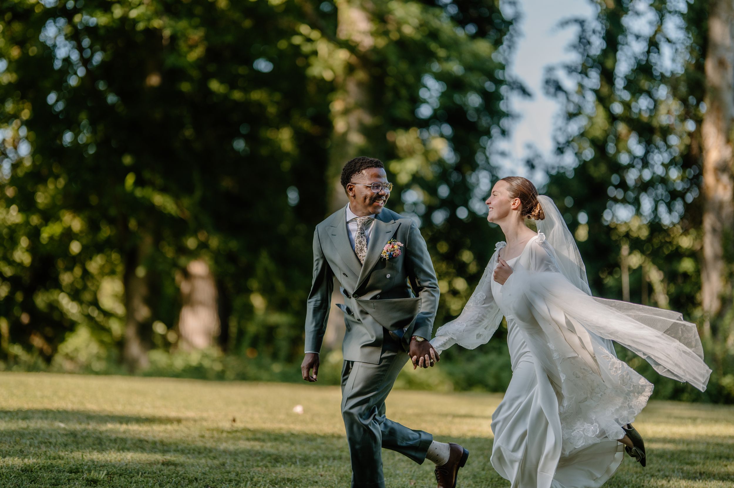 Joyful bride and groom running and laughing together on green lawn surrounded by tall trees on their wedding day.