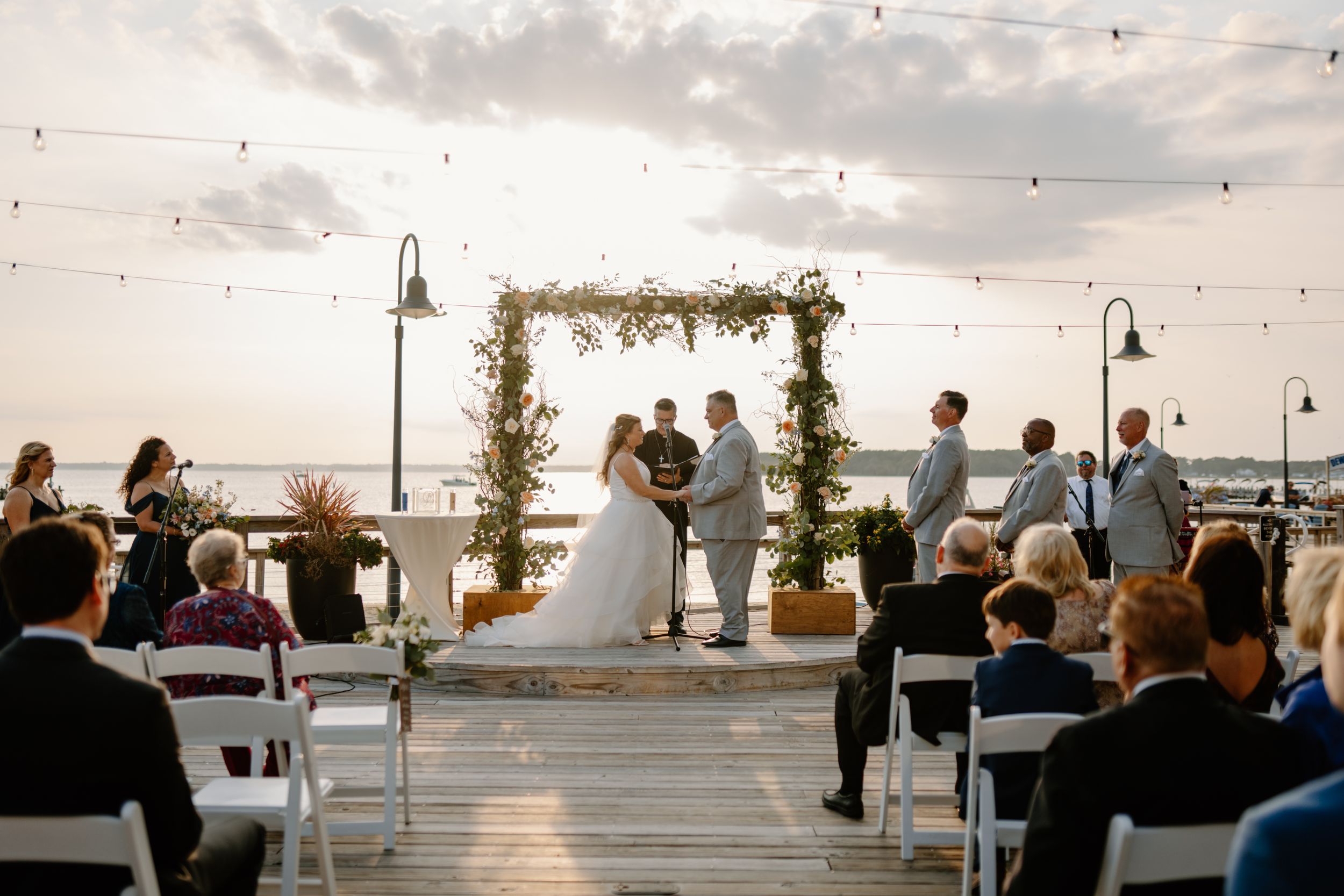 Sunset wedding ceremony on a waterfront deck with string lights and floral arch.