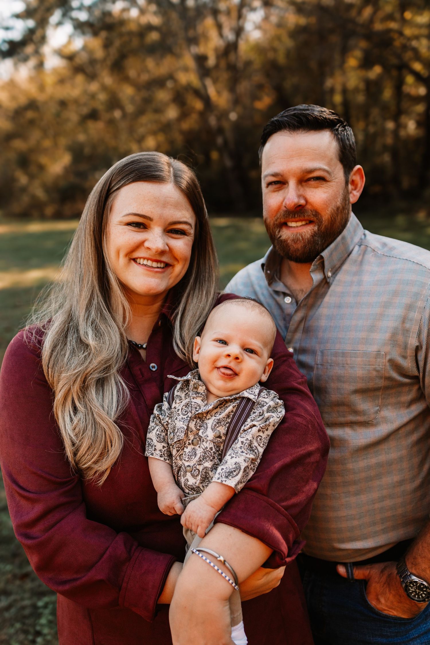A smiling woman and bearded man in autumn clothing pose outdoors with their baby during a family photo session in Knoxville, TN.