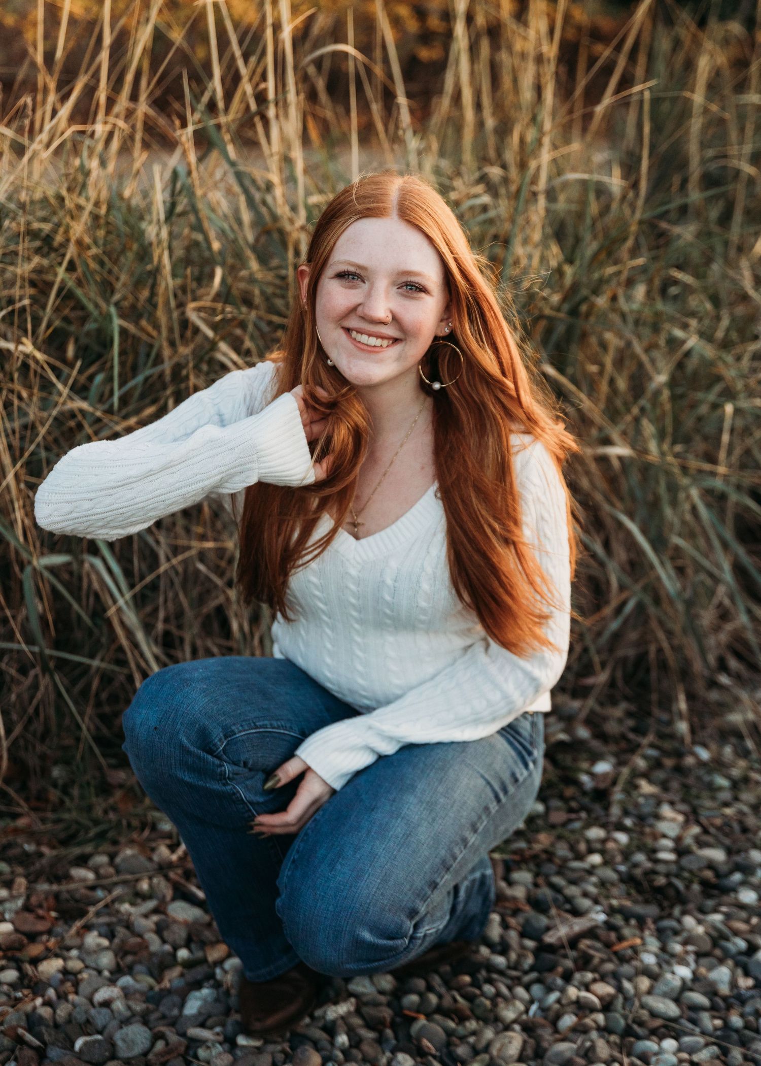 Smiling young woman with long red hair crouches on a pebble path in a white cable-knit sweater and jeans beside tall golden grass.