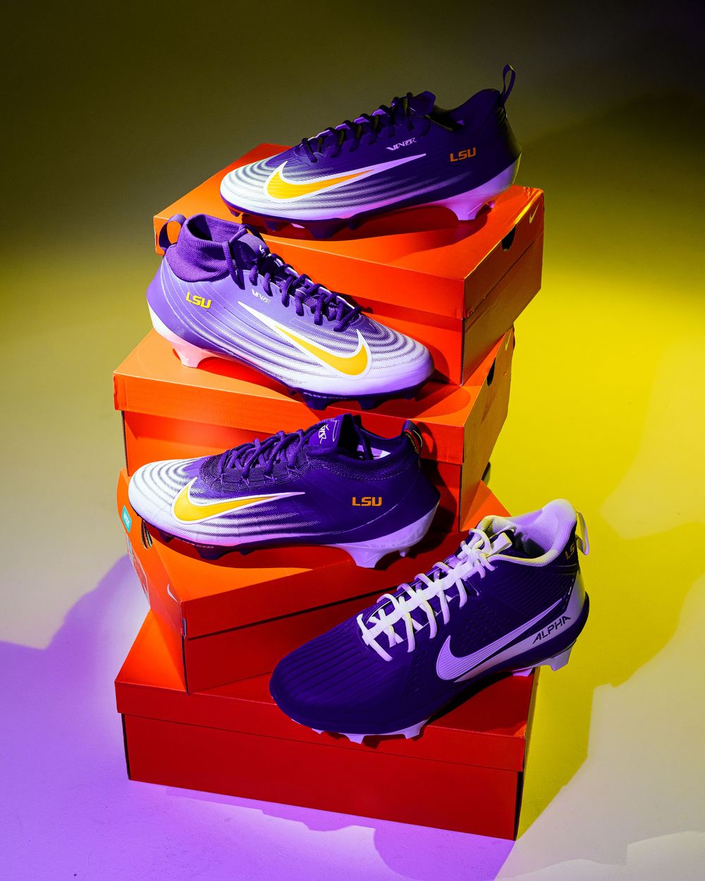 Four purple and gold LSU Nike baseball cleats displayed on stacked orange shoe boxes against a purple and yellow gradient backdrop.