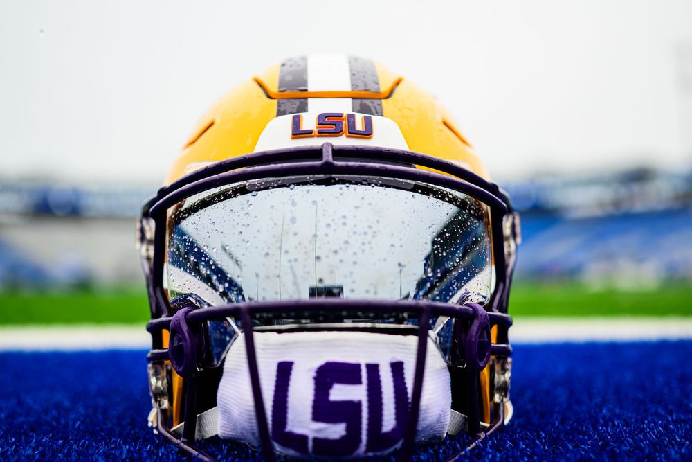 Gold LSU football helmet with purple and white striping and a rain-speckled visor rests on a blue turf field in a stadium.