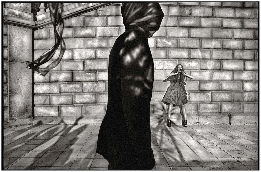 Black and white photo of a cloaked figure and a girl in a dramatic courtyard with stone walls and shadows.