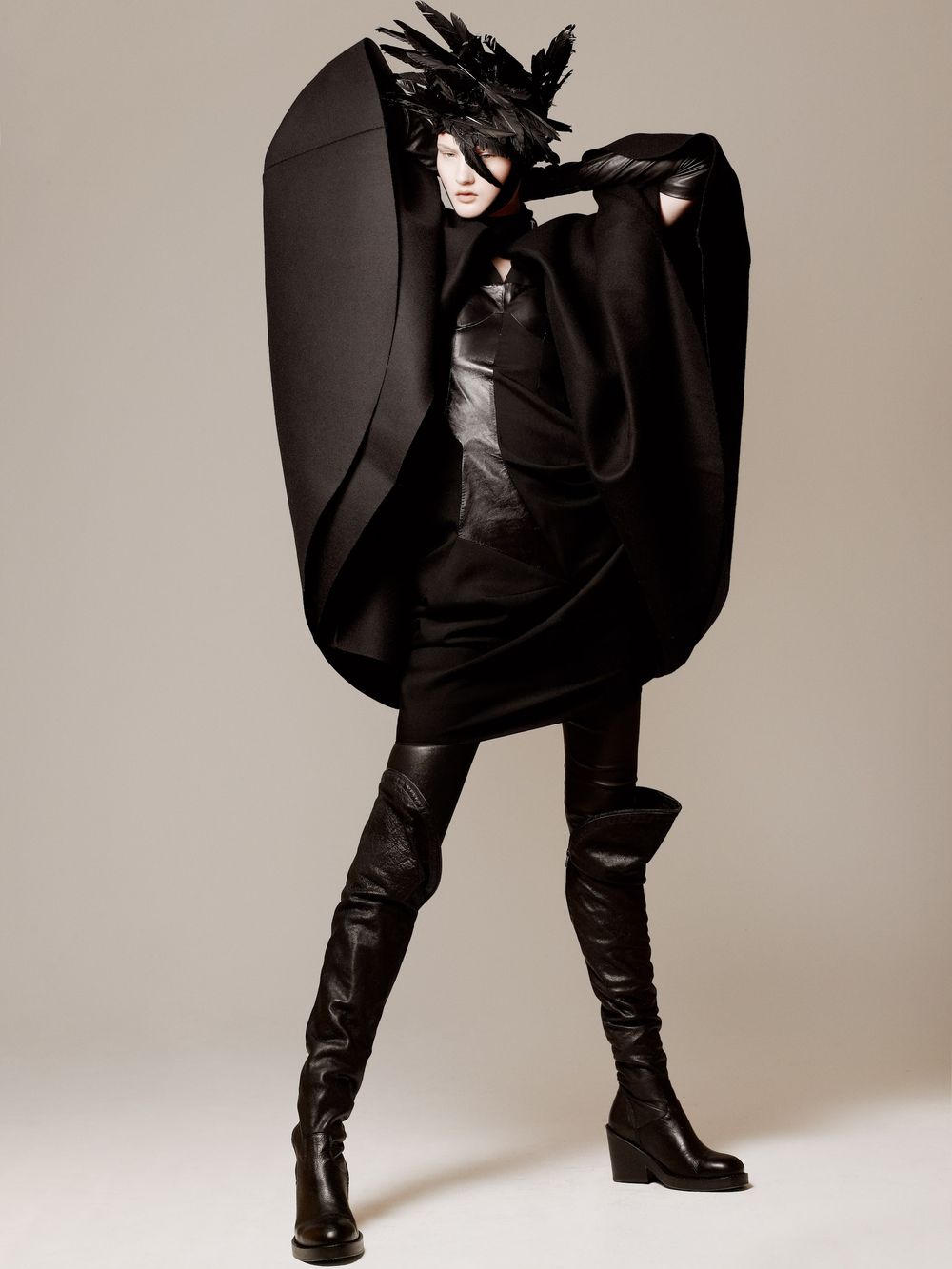 Model wearing dramatic all-black avant-garde fashion ensemble with oversized cape, thigh-high boots, and sculptural headpiece.