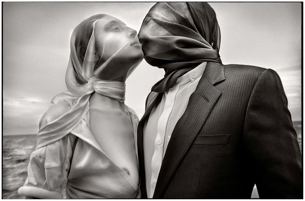 Black and white photo of a couple kissing with scarves blowing over their faces in the wind outdoors.