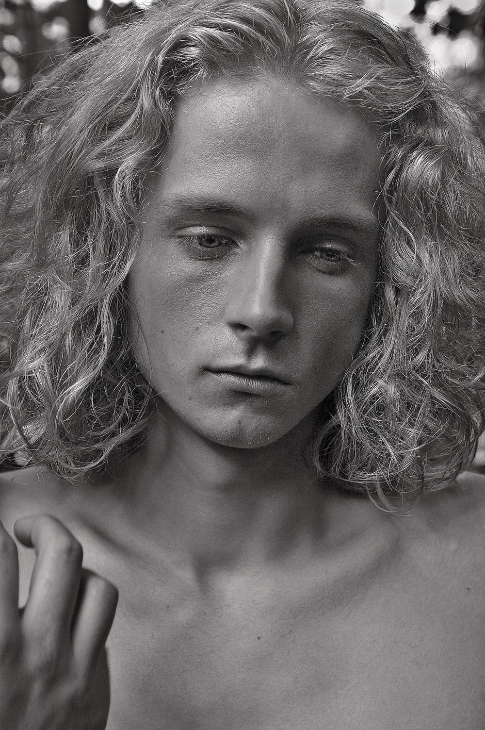 Black and white portrait of a young man with long curly hair, shirtless, gazing intensely at the camera outdoors.