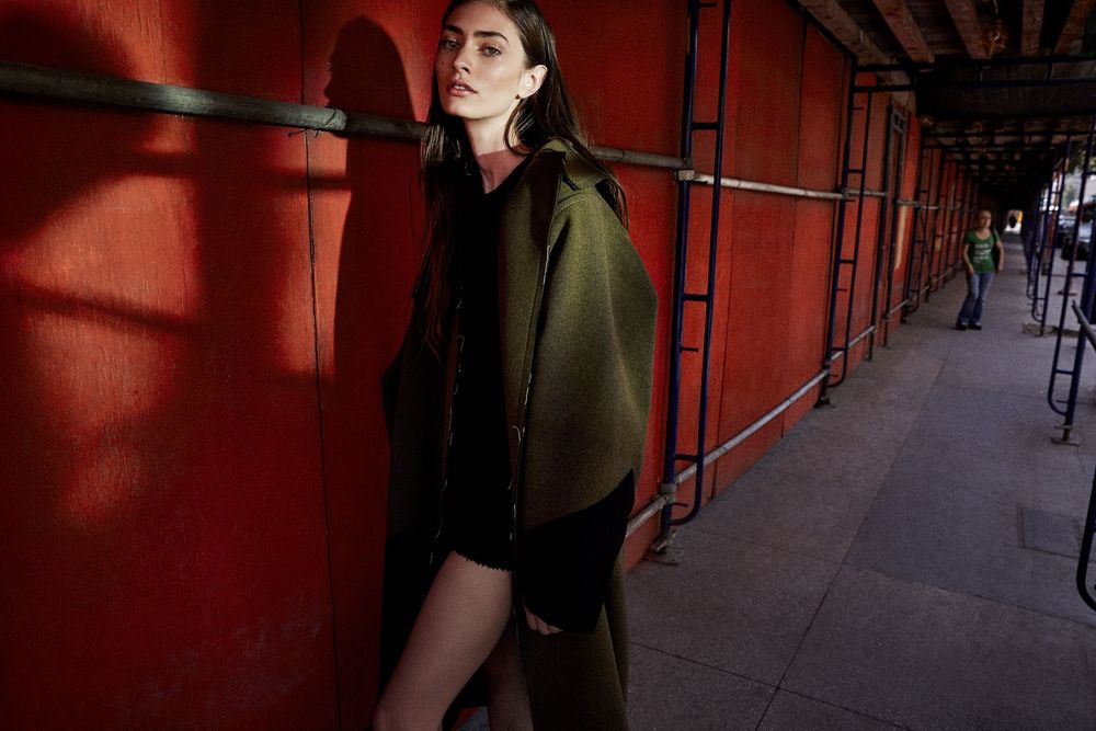 A model in an olive green oversized coat leans against a vibrant red wall in an urban setting.