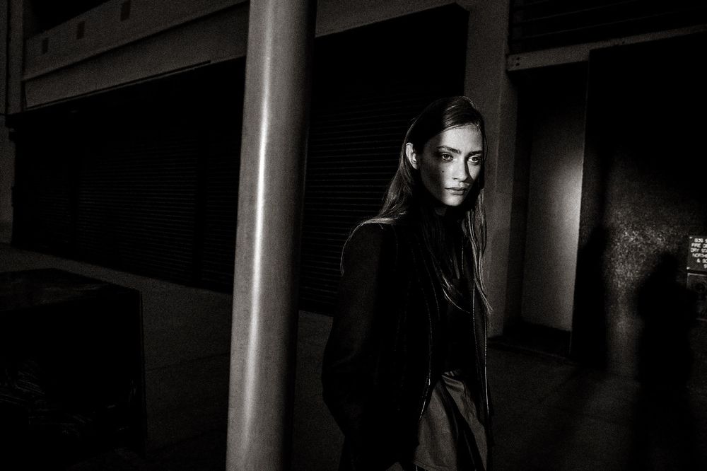 Young woman with long dark hair poses in black outfit against urban background in dramatic black and white photography.