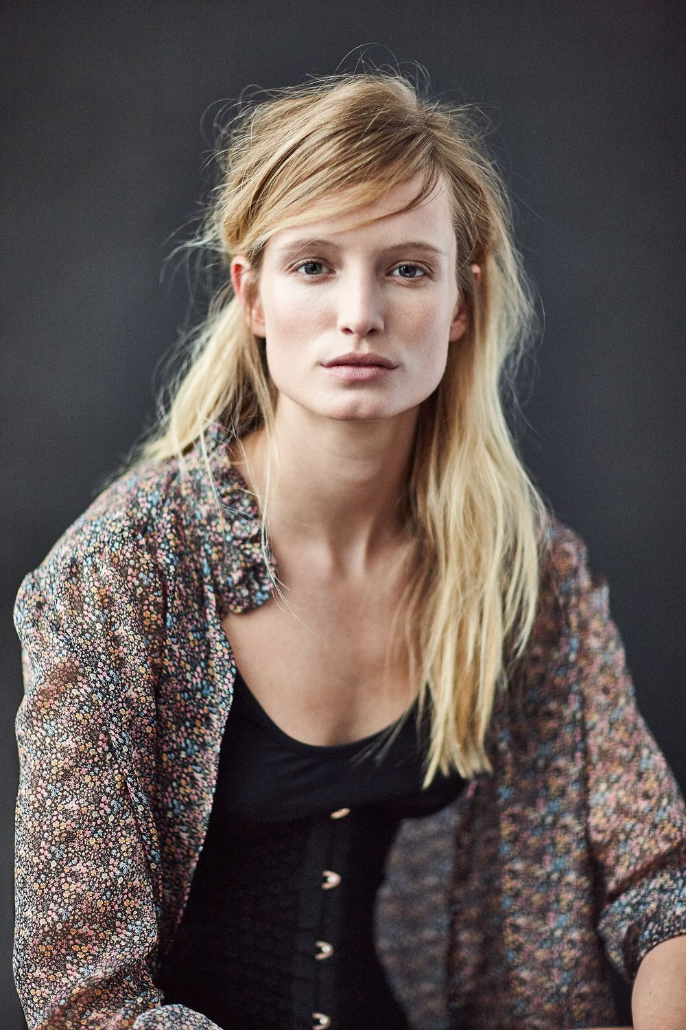 Young blonde woman in floral jacket and black top posing against dark gray background.