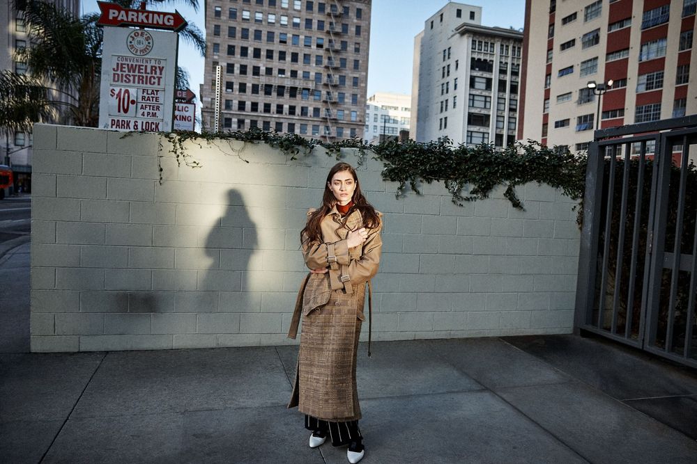 Woman in plaid trench coat stands on urban sidewalk with shadow on white brick wall and city buildings behind.