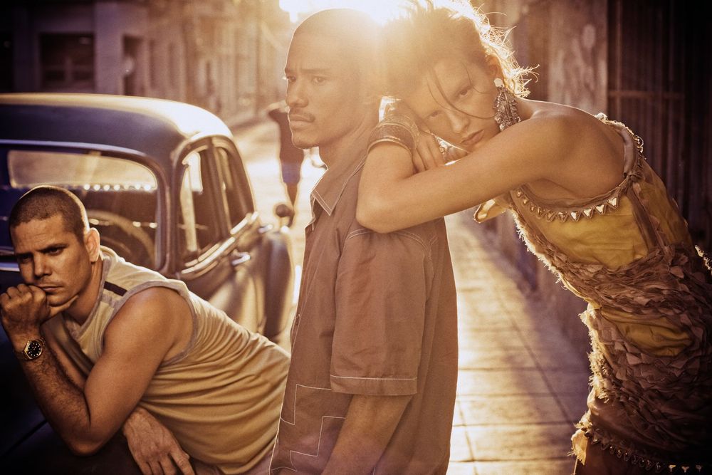 Three stylish people pose on a sun-drenched urban street beside a vintage car in warm golden light.