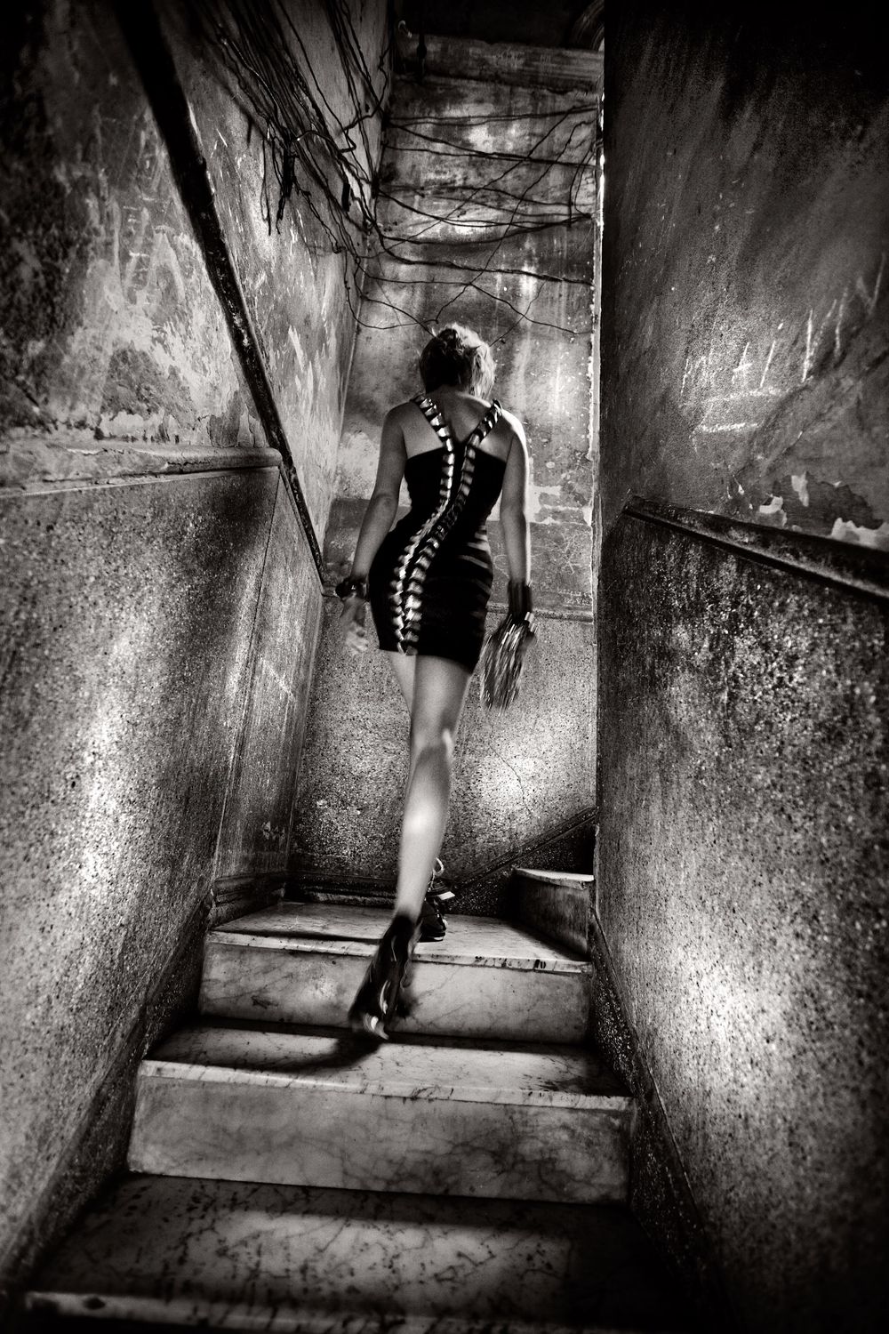 Woman in black dress ascending gritty urban stairwell, dramatic black and white photography with moody atmospheric lighting.