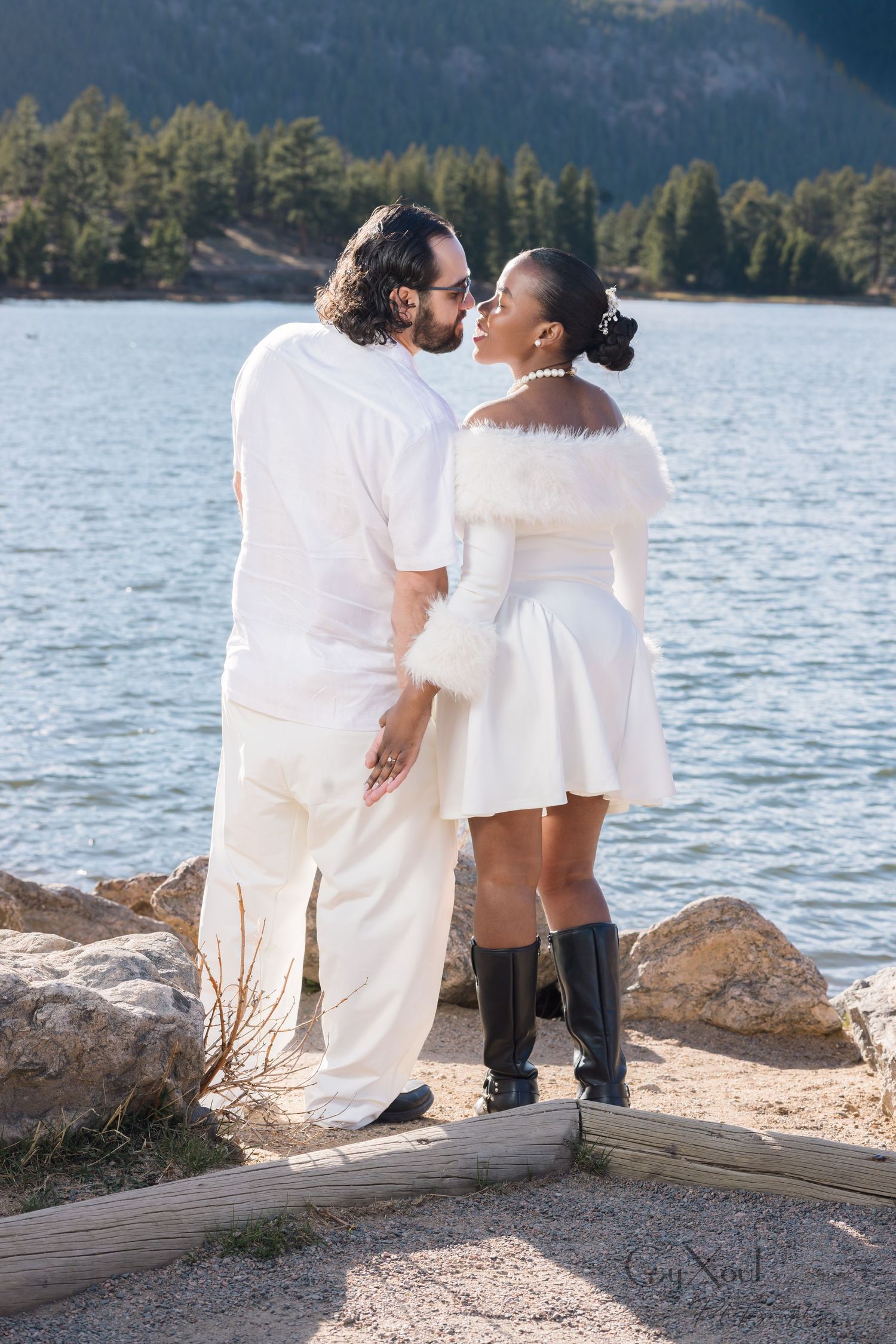 Couple in white outfits embracing by a mountain lake, the bride in a fur-trimmed dress and black boots, with pine trees in the background.