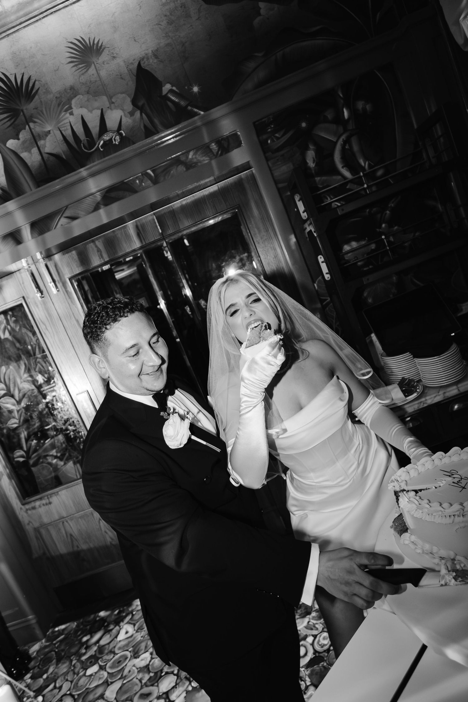 Black and white photo of a smiling bride in a veil and satin gloves cutting a wedding cake with her groom in a tuxedo.