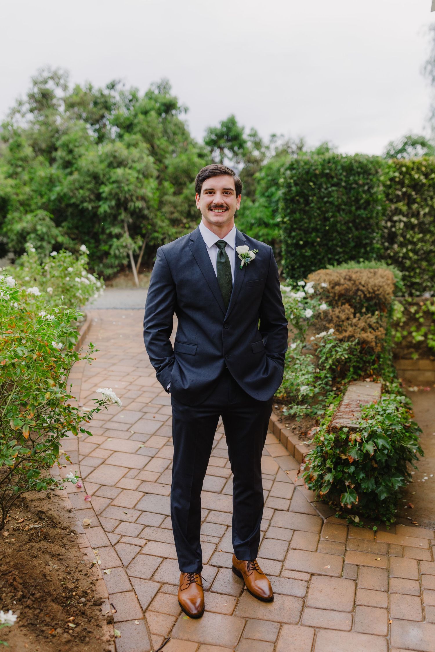 Groom with a mustache wears a navy suit, green tie and boutonniere while standing on a brick garden path.