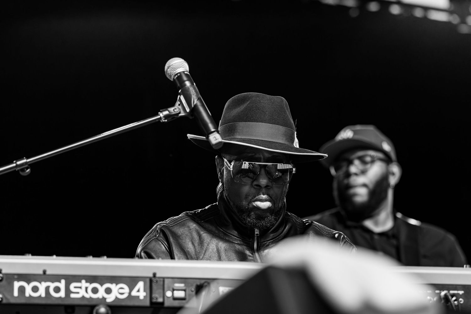 Black and white photo of a musician in a hat and leather jacket performing at a Nord Stage 4 keyboard on stage.
