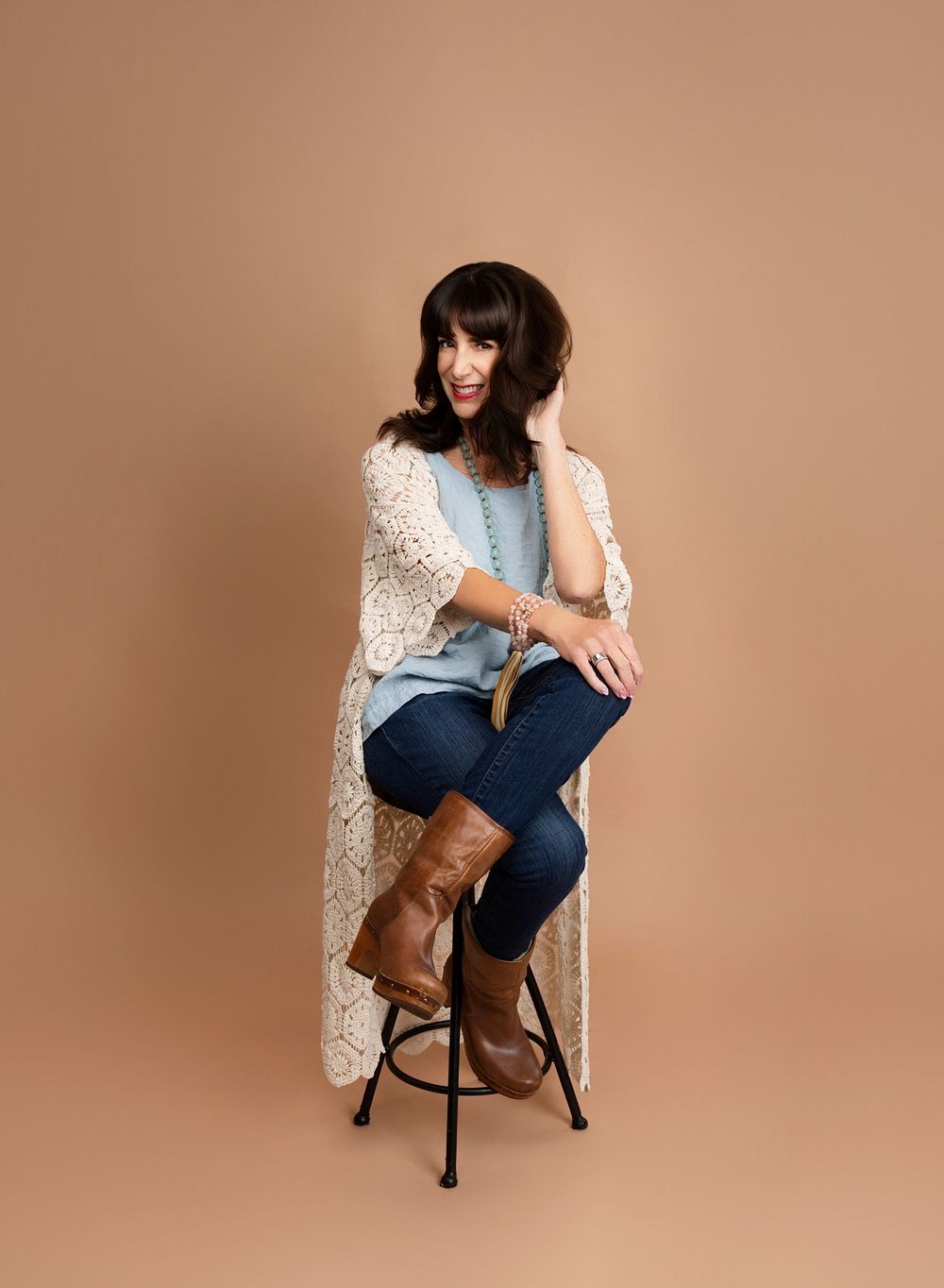 Woman in a black top and jeans seated on a stool against a tan backdrop, personal branding portrait