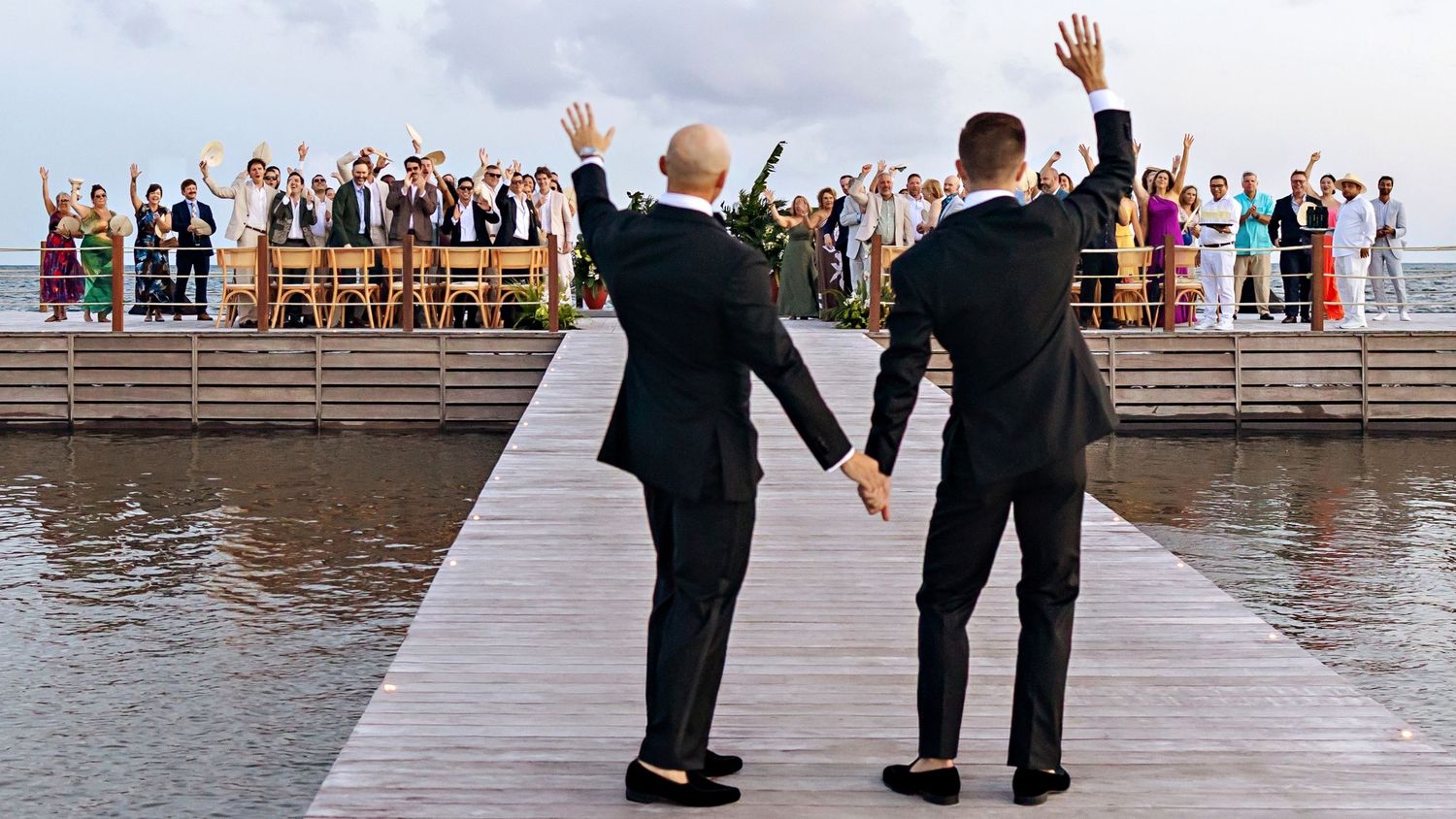 Two grooms in black tuxedos wave to wedding guests on a waterfront dock during an outdoor ceremony.