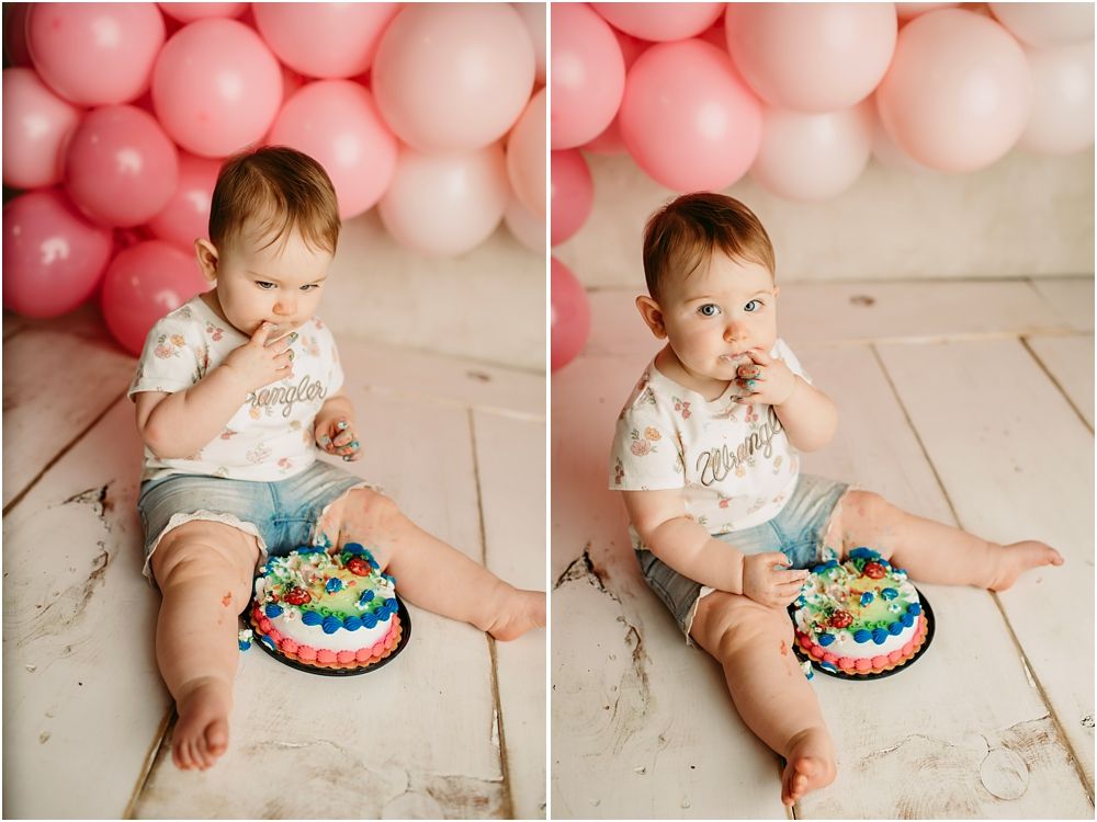 Baby girl enjoys her first birthday cake smash session with pink balloons backdrop on white wood floor.