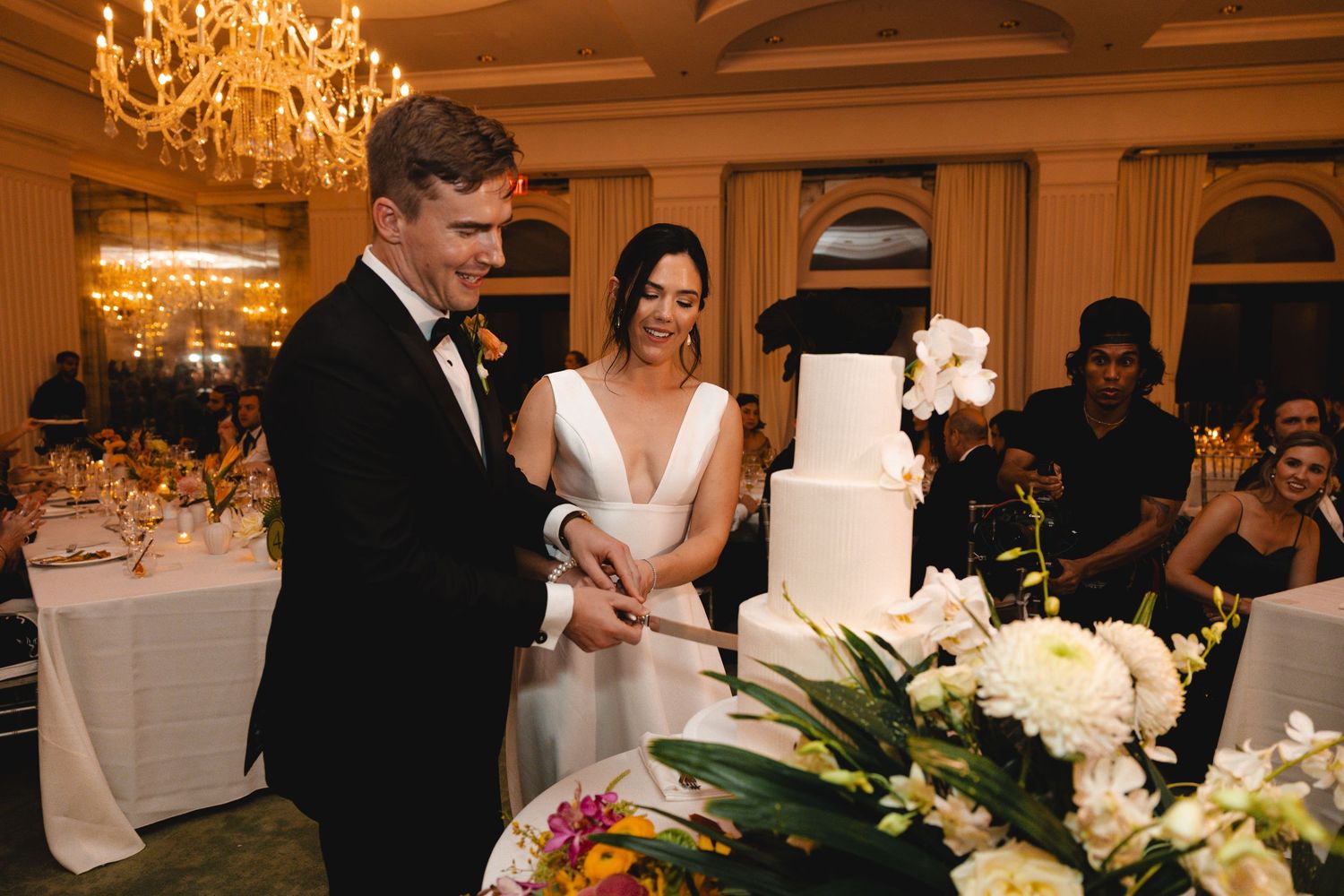 Bride and groom share a kiss beside a white wedding cake in an elegant reception hall with chandeliers and seated guests.