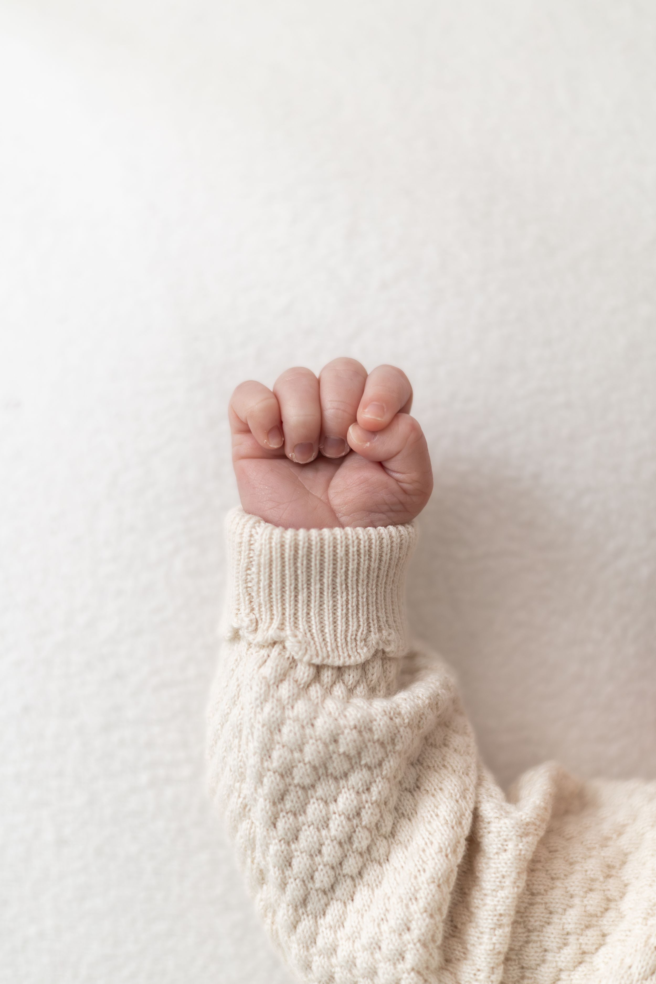 Close-up of a newborn baby's tiny clenched fist in a cream knit onesie against white background.