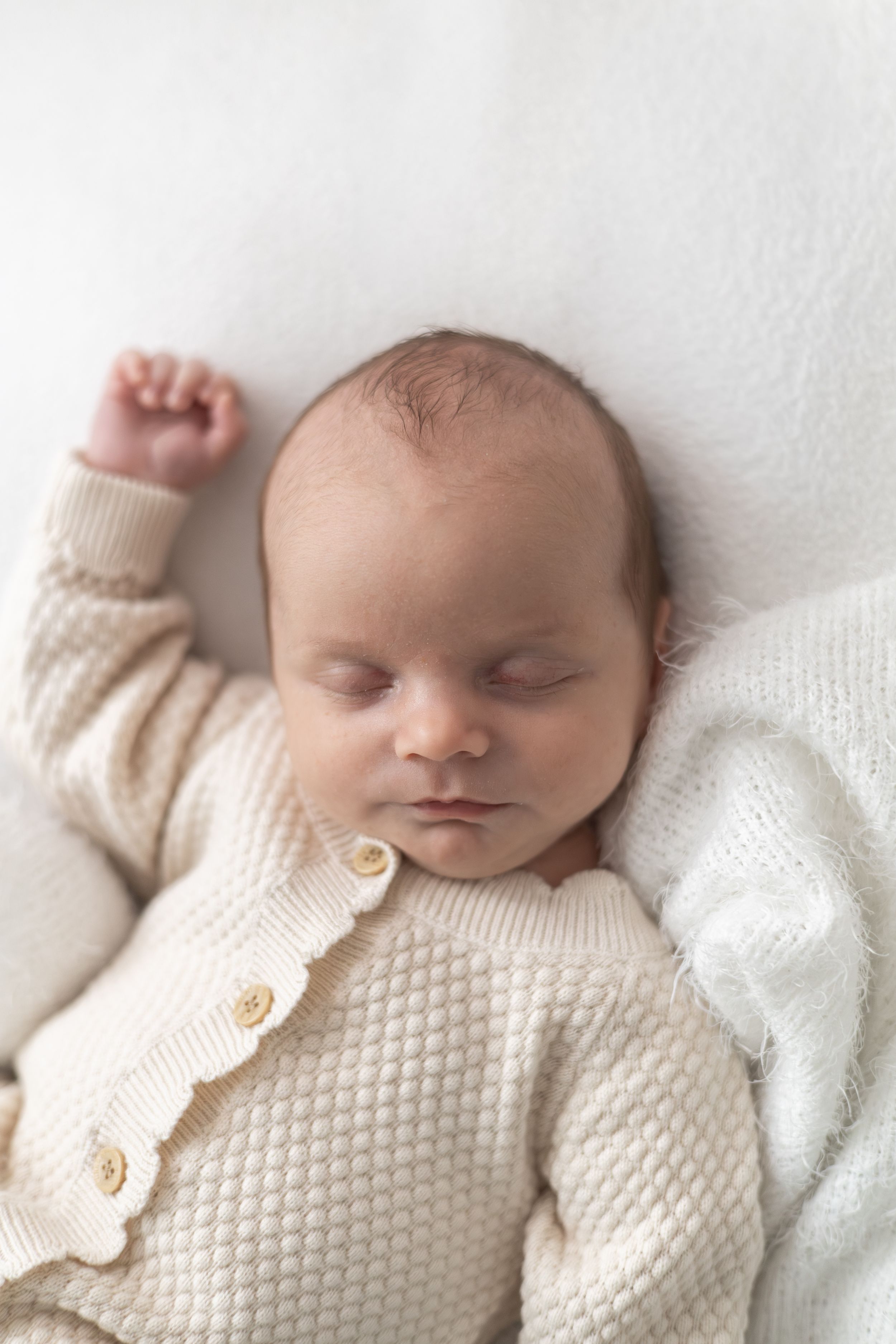 Sleeping newborn baby in cream knit cardigan with gold buttons, resting peacefully on white blanket.