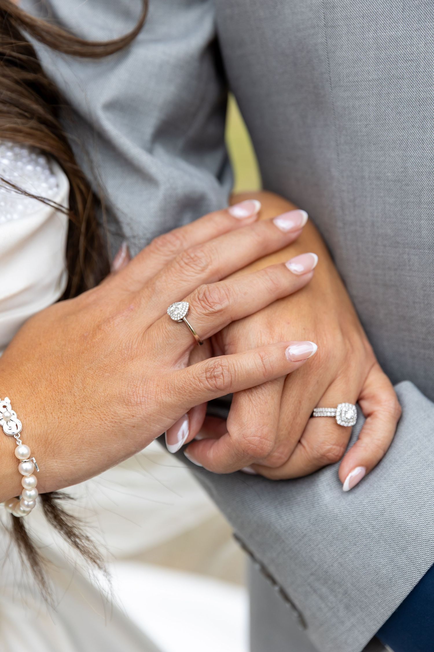 Close-up of a bride and groom holding hands, showing sparkling diamond engagement and wedding rings and a pearl bracelet.