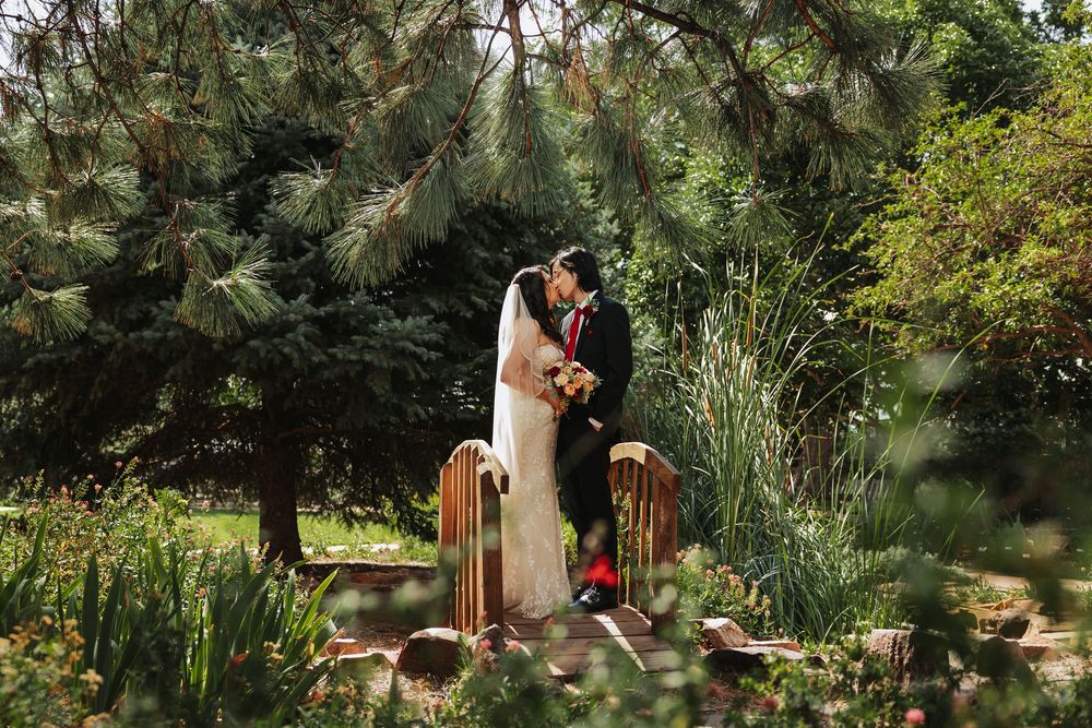 Bride and groom share a romantic kiss on a garden bridge surrounded by lush green trees on their wedding day.