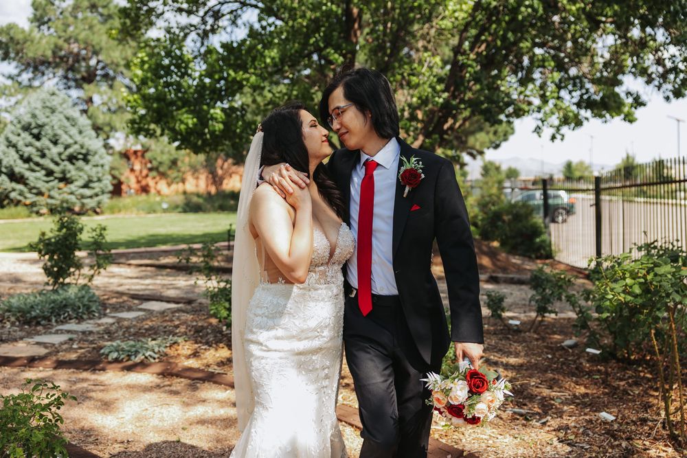 Asian bride and groom share romantic moment walking in garden, bride in white gown with veil, groom in black suit with red tie.