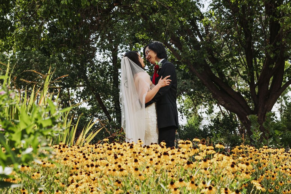 Bride and groom embrace among vibrant yellow flowers in a lush garden, bride's veil flowing gracefully.