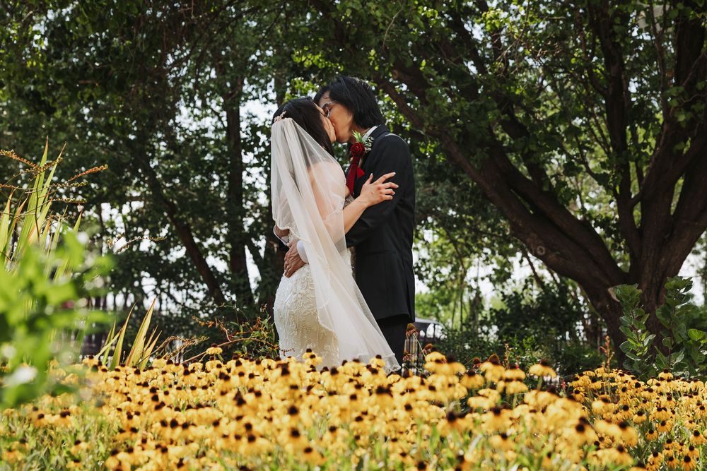 Bride and groom kissing in a lush garden surrounded by vibrant yellow flowers on their wedding day.