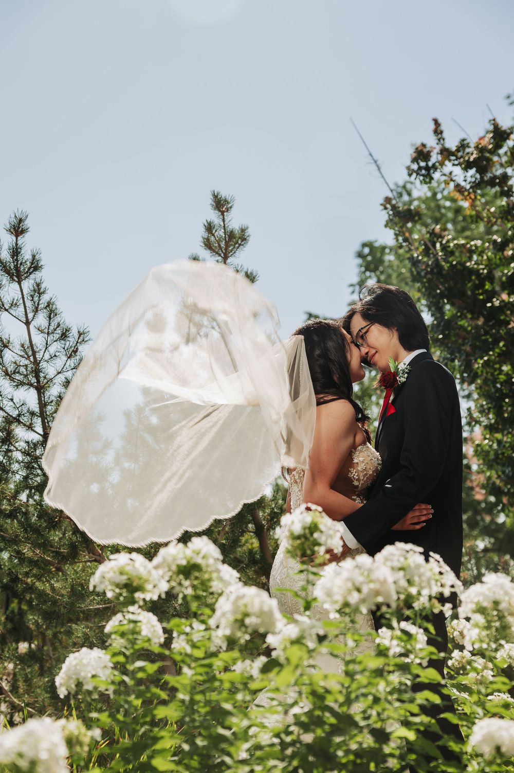 Bride and groom share a romantic moment outdoors, with veil flowing in wind, surrounded by white flowers and green trees.