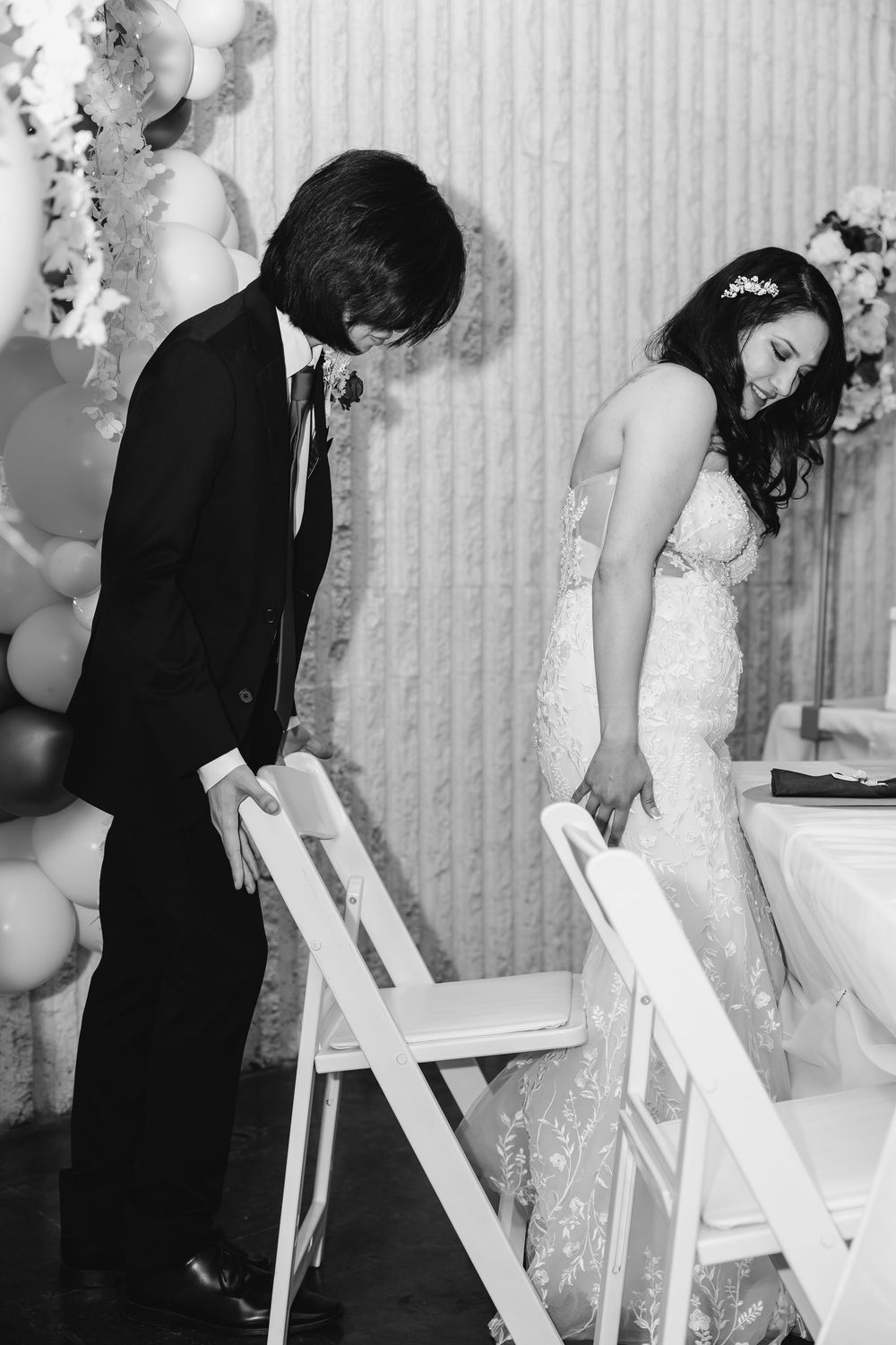 Black and white photo of bride in lace gown and groom in suit laughing together at wedding reception.