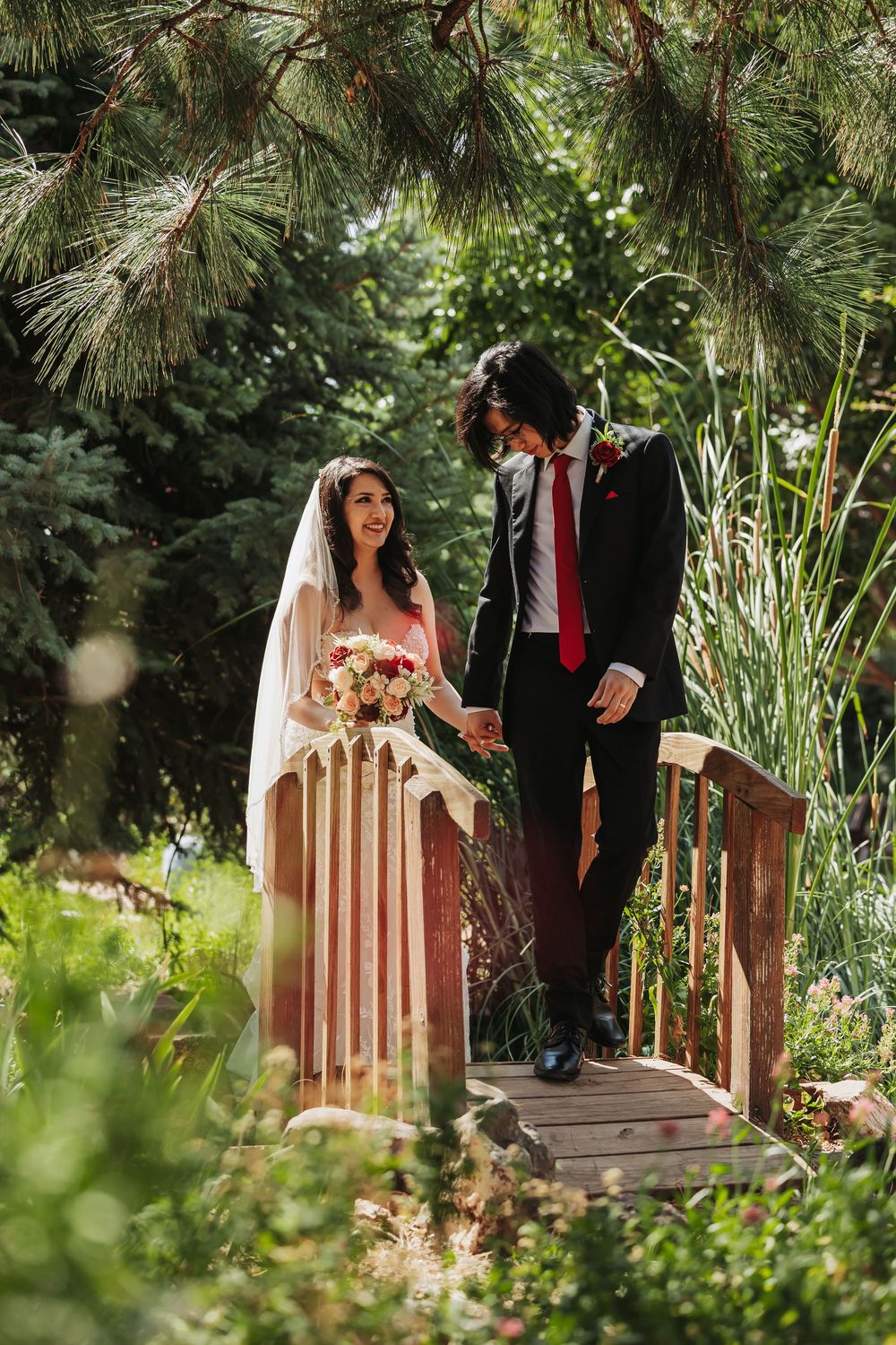 Bride and groom holding hands on a wooden garden bridge, surrounded by lush greenery on their wedding day.