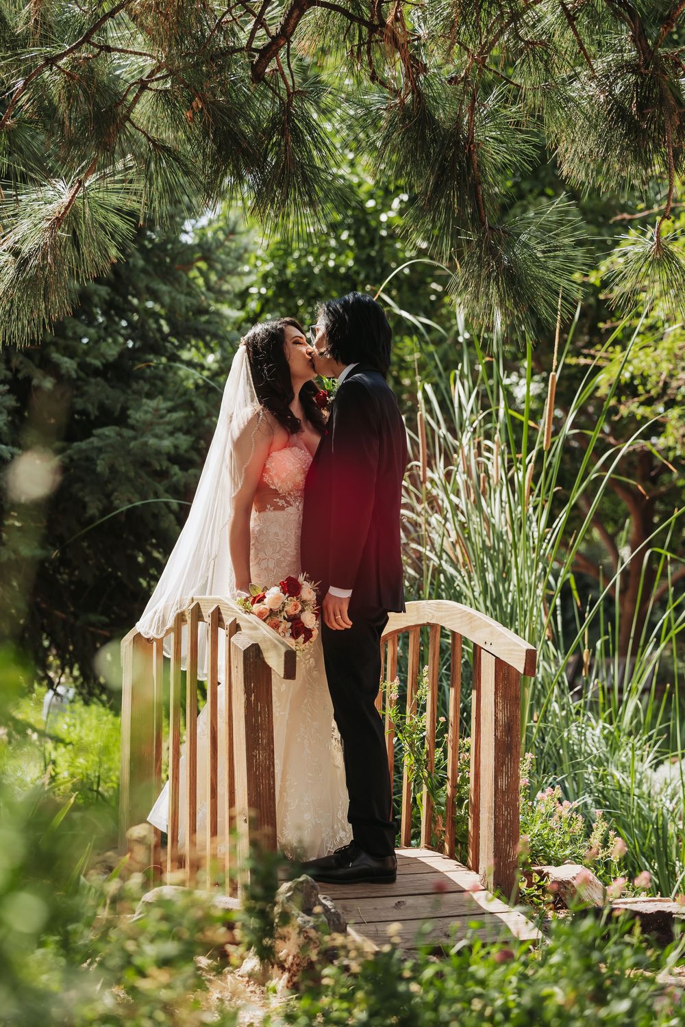 Bride and groom share a kiss on a wooden bridge surrounded by lush greenery, bride holding a bouquet in a white gown.