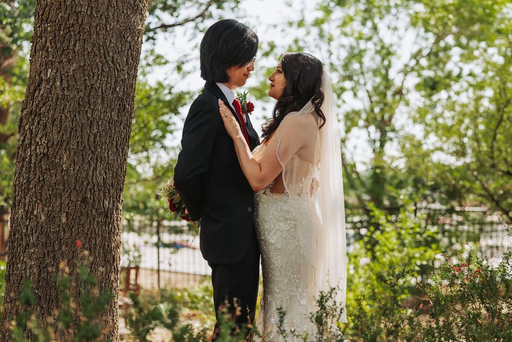 Bride and groom share a romantic moment near a tree, bride in lace gown and groom in black suit outdoors.