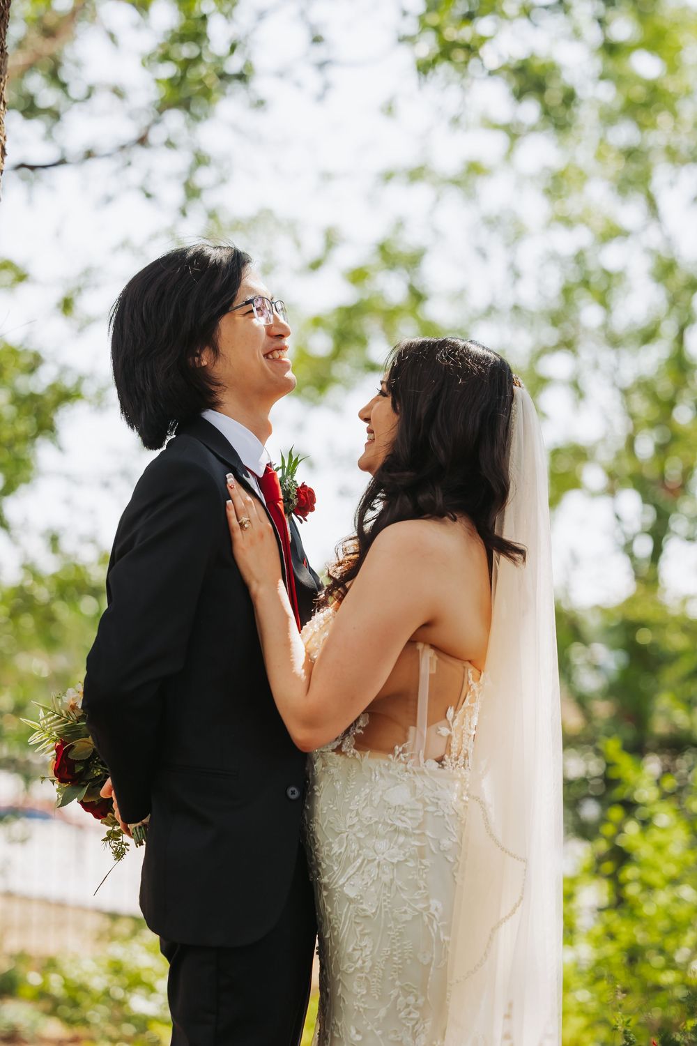 A bride and groom share a joyful moment outdoors, smiling at each other, with the bride in a lace gown and veil.