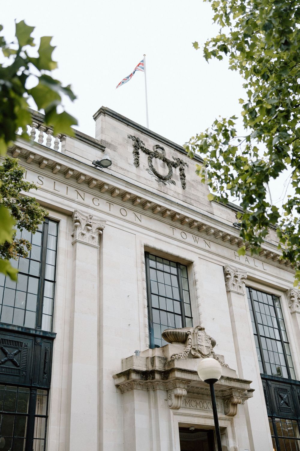 Islington Town Hall exterior with white neoclassical facade, British flag on rooftop, and green trees framing the building.