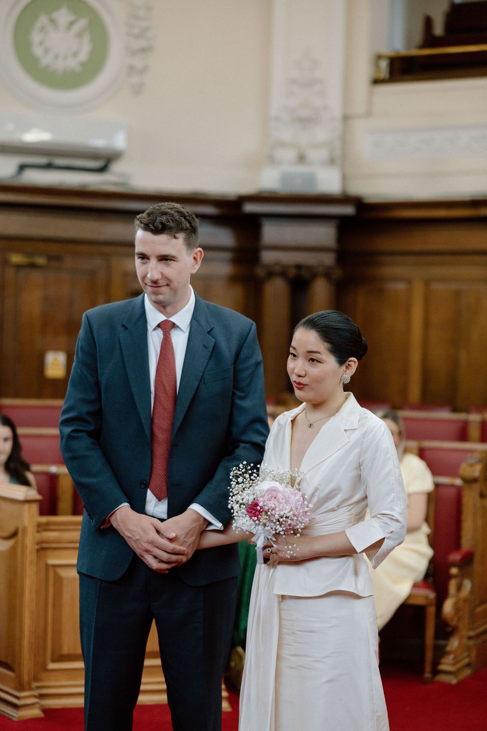 Couple stands at civil wedding ceremony, groom in dark suit with red tie, bride in white outfit holding pink bouquet.